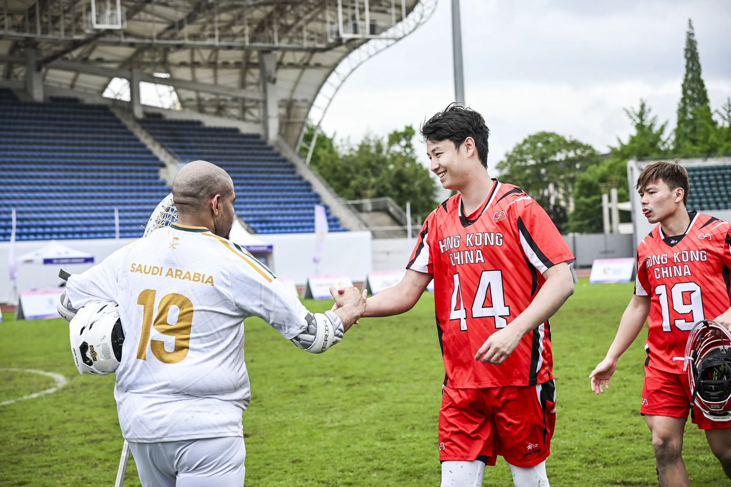 Two Asian men in sports uniforms shaking hands after a game, one wearing a red uniform with 'Hong Kong China' and the other a white jersey with 'Saudi Arabia', on a sports field with empty stadium seats in the background.