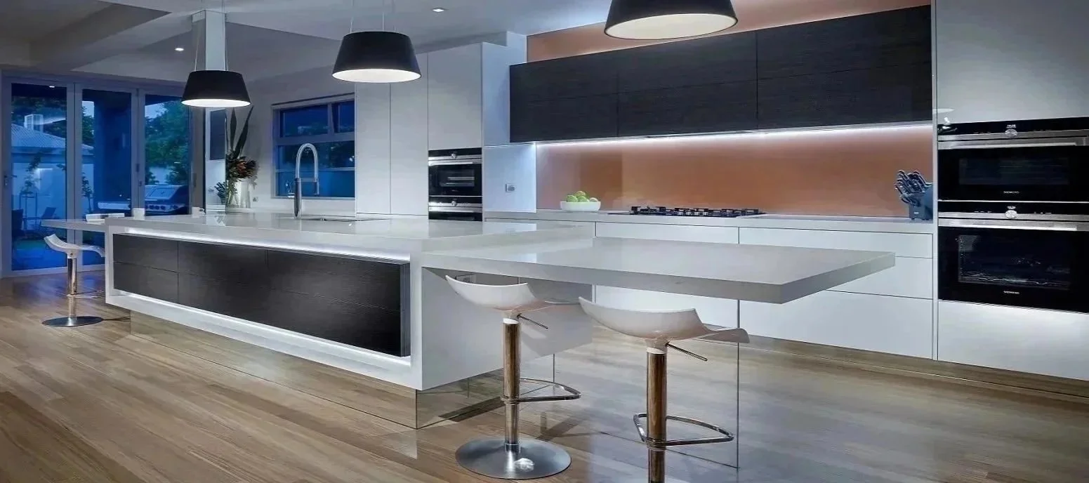 Modern kitchen with white countertops, black and wooden cabinets, a large island with a white surface, two white barstools, pendant lights, and built-in ovens.