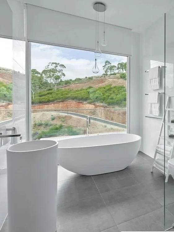 Modern bathroom with a freestanding bathtub, a floor-standing sink, and a large window overlooking a hillside with greenery.