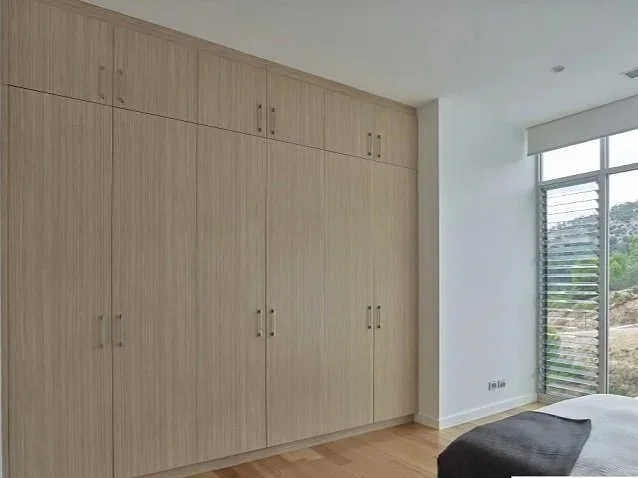 Modern bedroom with large built-in wooden wardrobe and a window with horizontal blinds overlooking greenery.