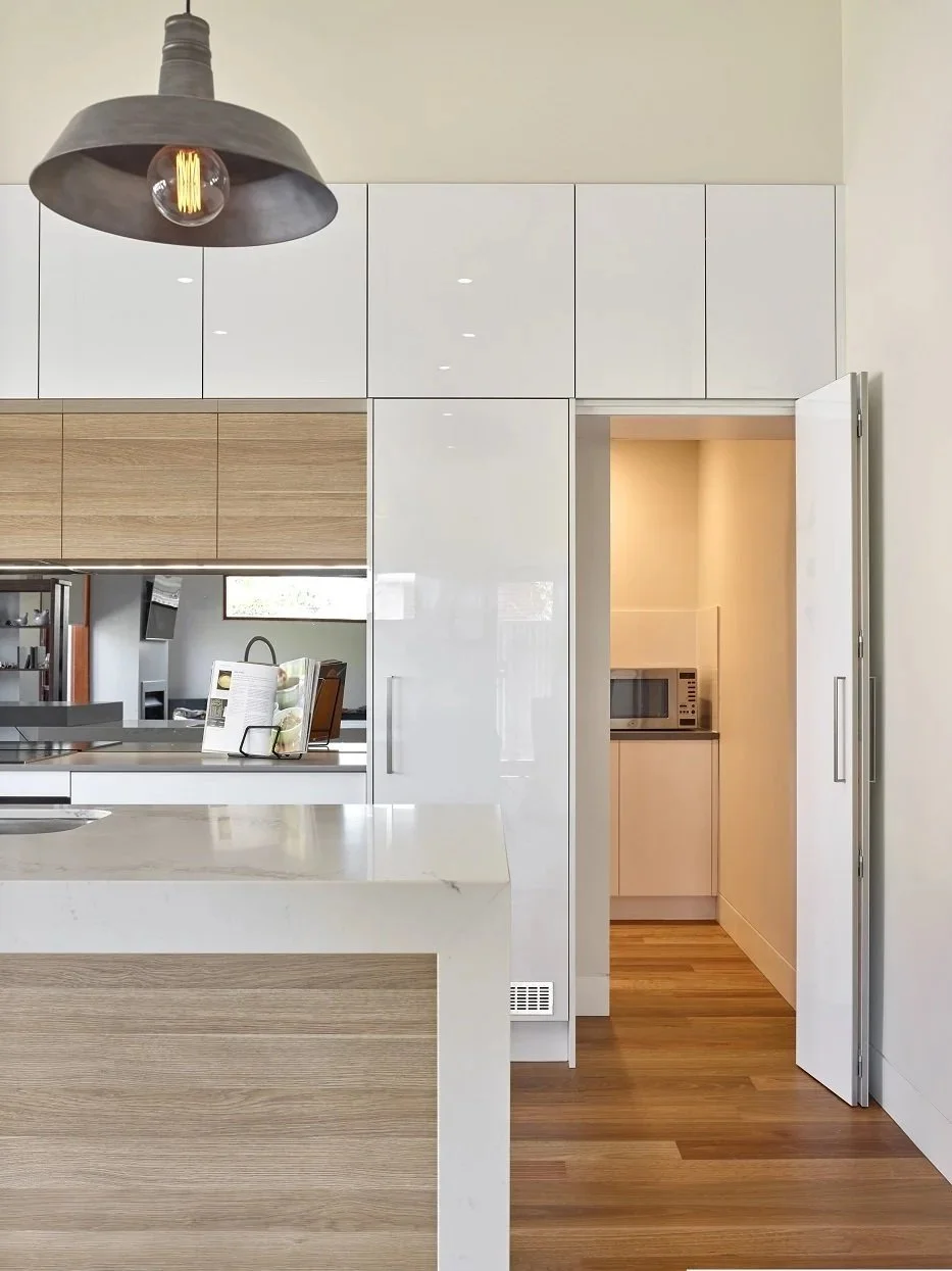 Modern kitchen with white cabinets, a wooden accent wall, and a small pantry with a microwave. A gray industrial pendant light hangs above the kitchen island.