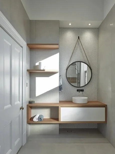 Modern bathroom vanity with a round mirror hanging from a leather strap, a vessel sink, wooden countertop, and shelves with towels, in a minimalist design.