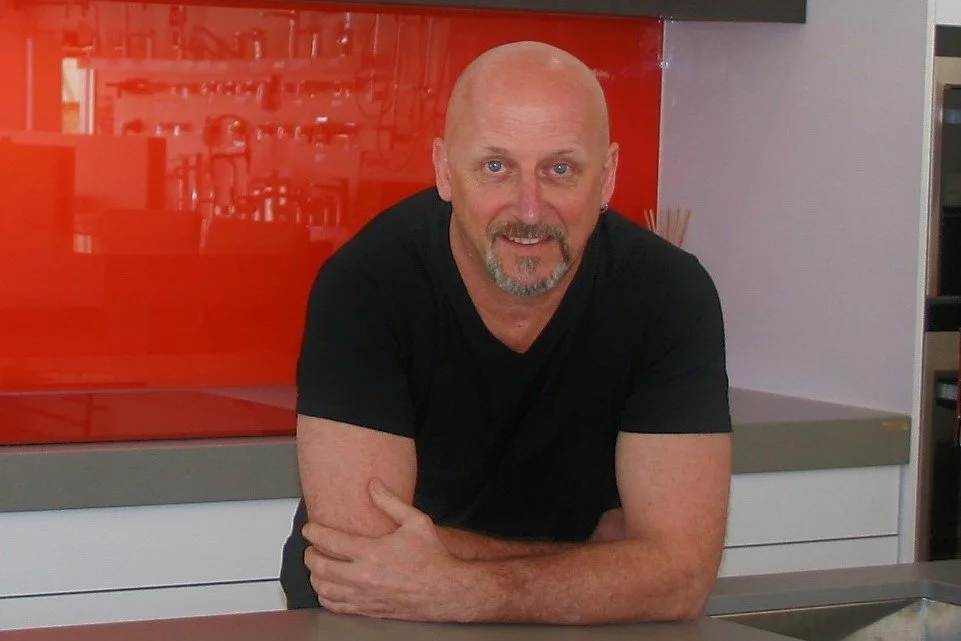 A middle-aged man with a bald head and goatee, wearing a black t-shirt, leaning on a counter in a kitchen with a red backsplash.