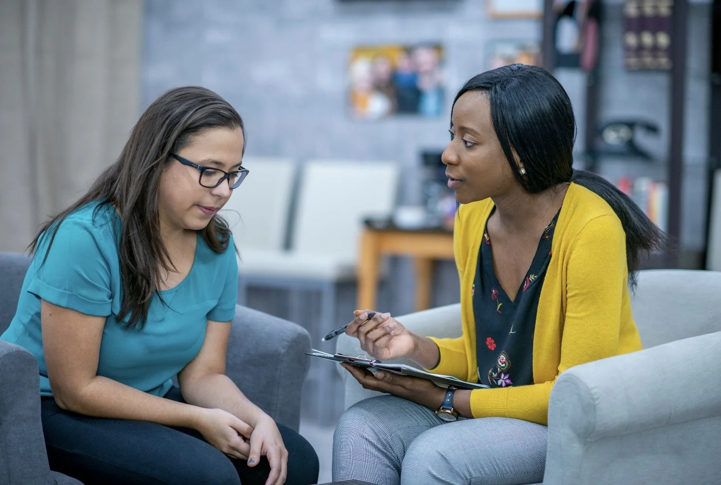 Two women seated in a professional office setting, engaged in a parent consultation session. The woman on the right is a therapist holding a notebook and pen, while the woman on the left is the mother and is listening attentively.
