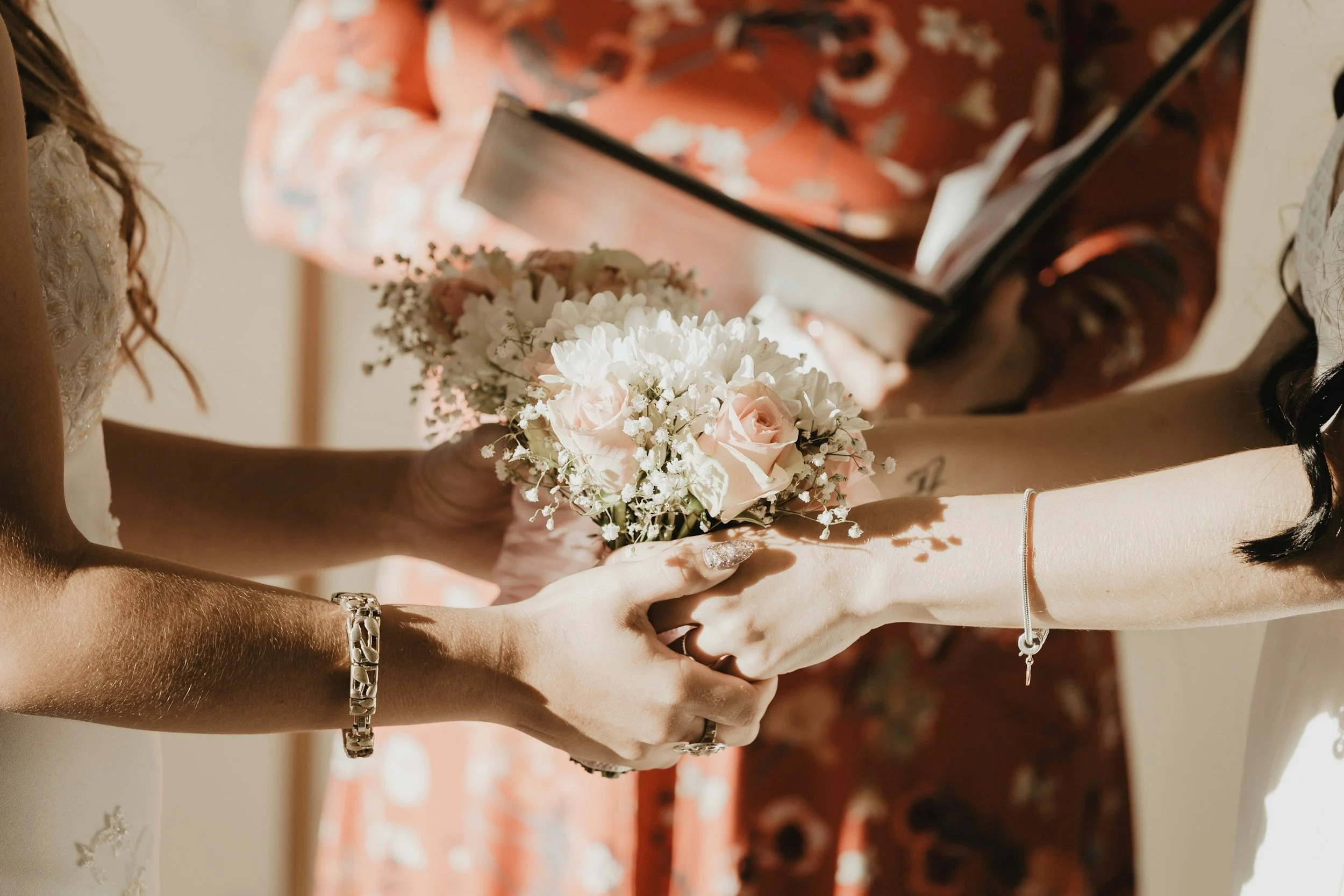Two women exchanging wedding vows, with one woman holding a bouquet of white and pink flowers, and the other woman reaching out.
