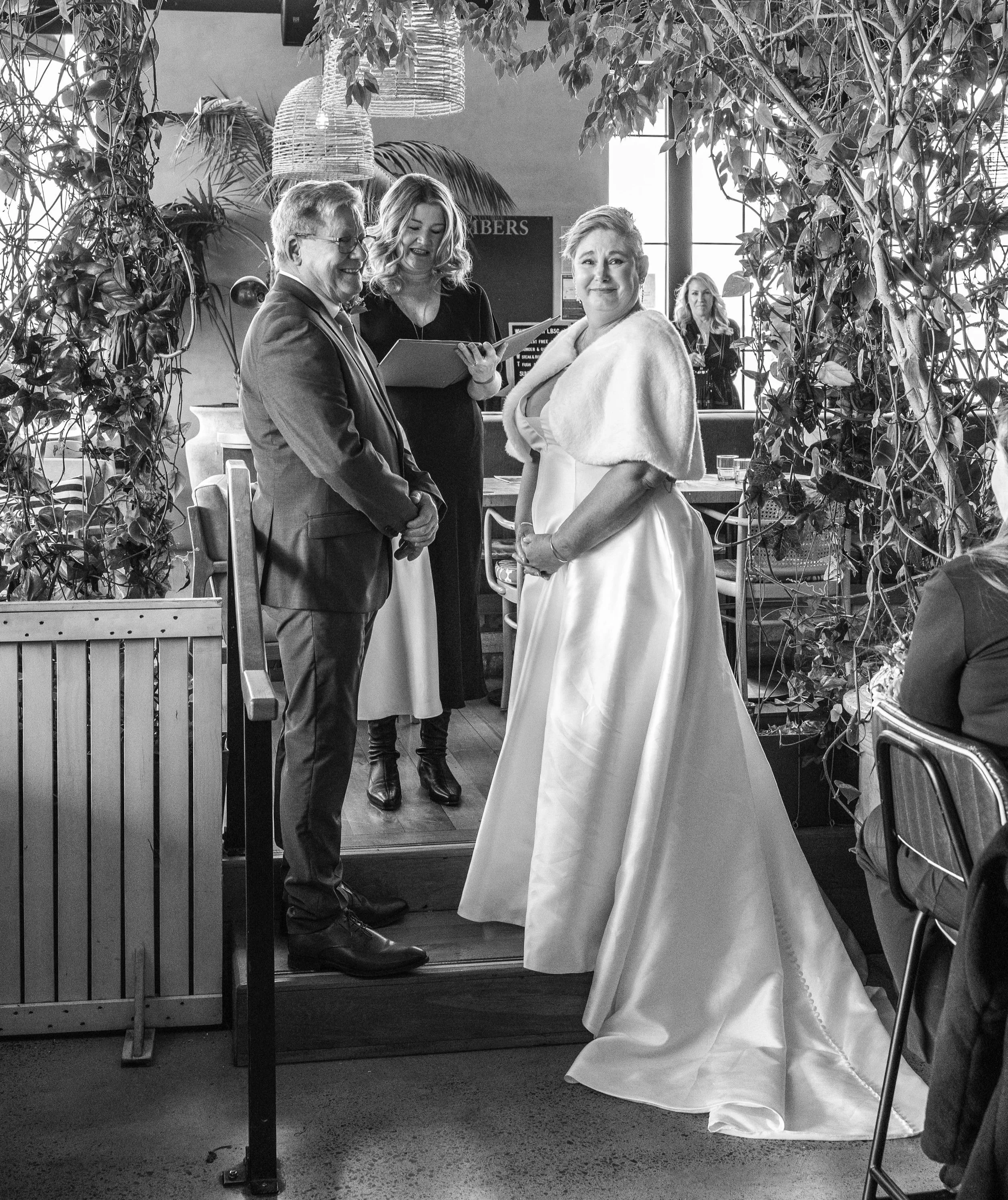 A bride in a white wedding dress and cape stands inside an art gallery or restaurant, speaking with a man in a suit. Behind her, a woman holds a notepad, observing and smiling. The scene is decorated with plants and hanging light fixtures.