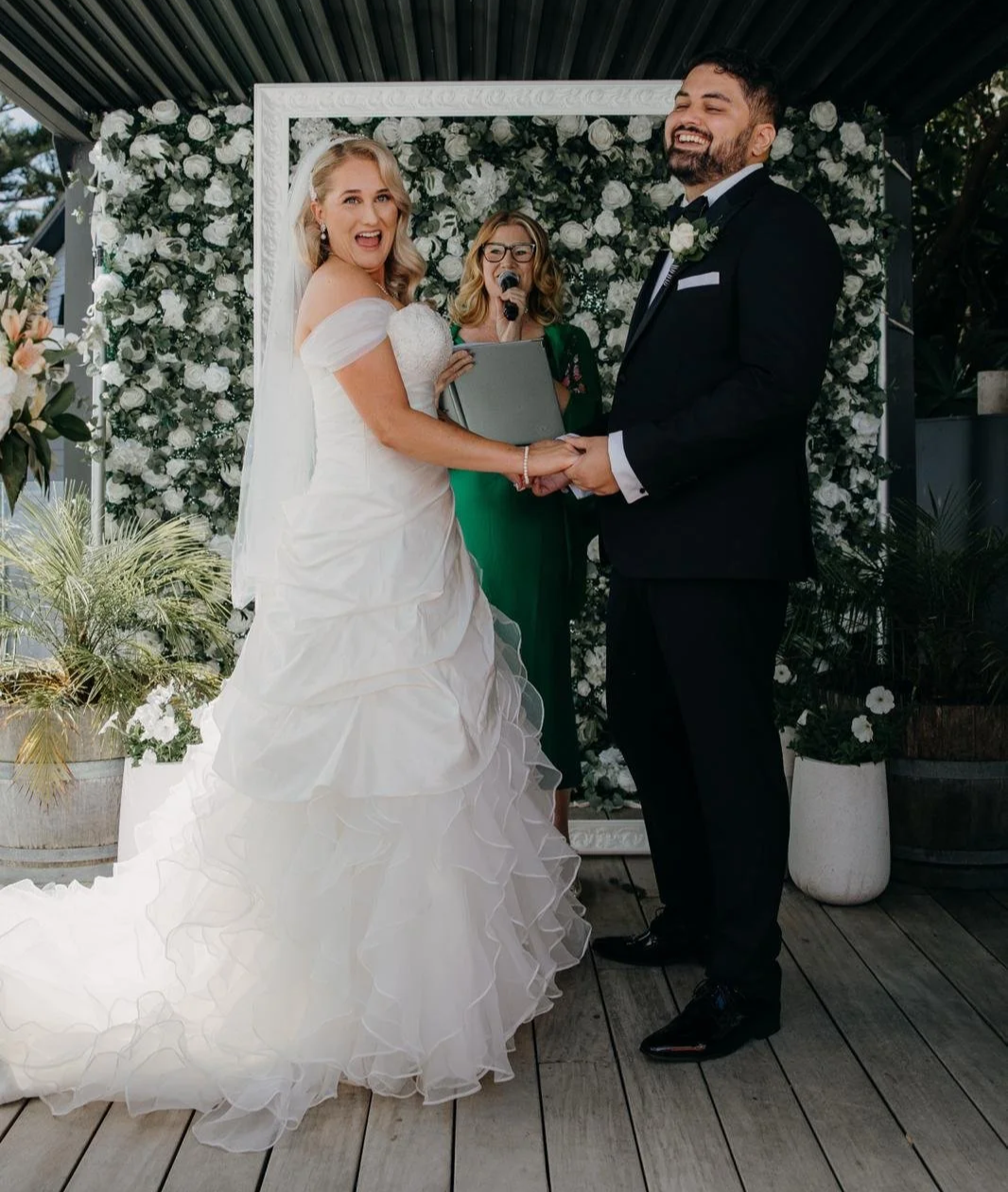 Bride and groom holding hands during their wedding ceremony, with officiant in the background, surrounded by white flowers and greenery.
