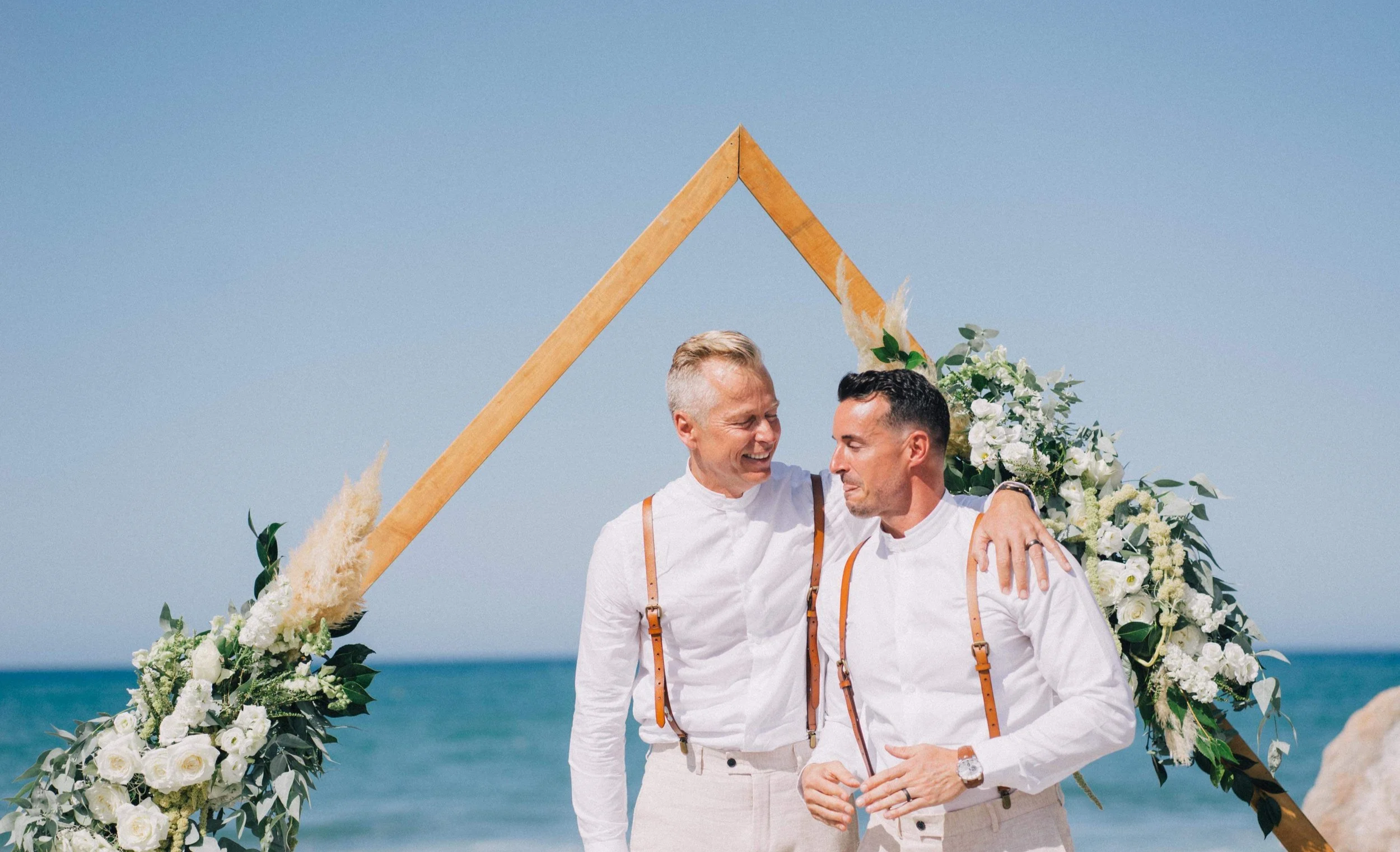Two men, dressed in white shirts and suspenders, embracing and smiling at each other during a wedding ceremony on the beach, with a floral arch and blue sky in the background.