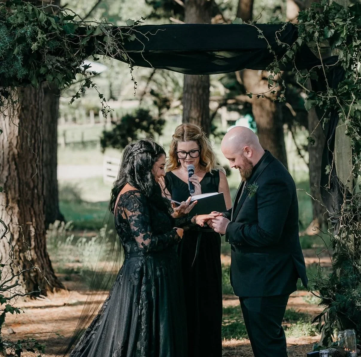 A couple getting married outdoors under a decorated arch, with a woman officiant reading vows and holding a microphone during a wedding ceremony.