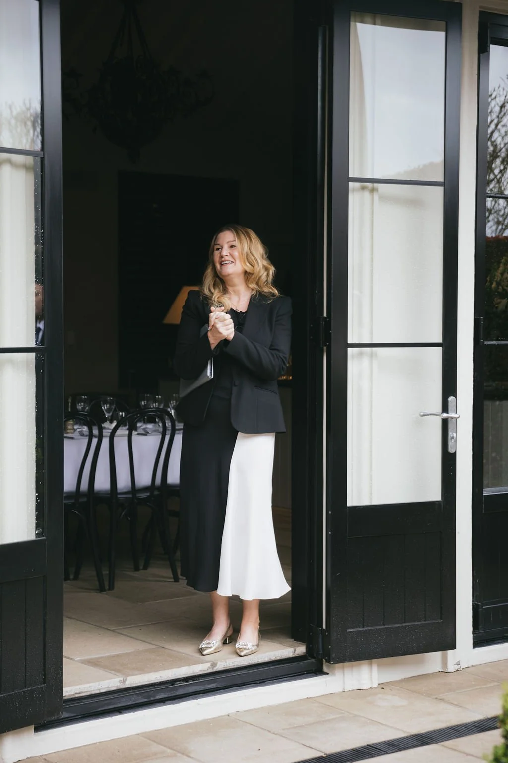 A woman in a black blazer and a black-and-white dress stands in an open doorway, smiling and clapping her hands.