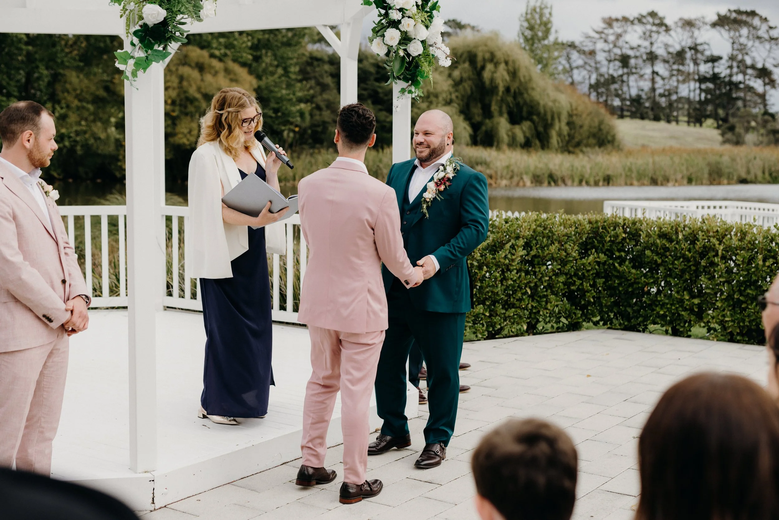 A wedding ceremony taking place outdoors by a lake, with a couple holding hands and exchanging vows under a white floral arch, surrounded by officiants and guests.
