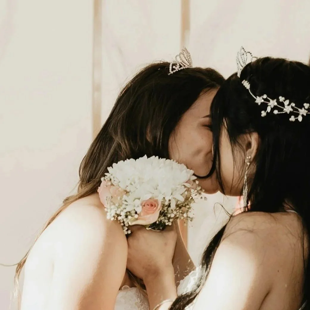Two women wearing tiaras share a kiss at a wedding, one holding a bouquet of flowers.