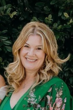 A woman with blonde hair smiling in front of green foliage, wearing a green floral blouse.