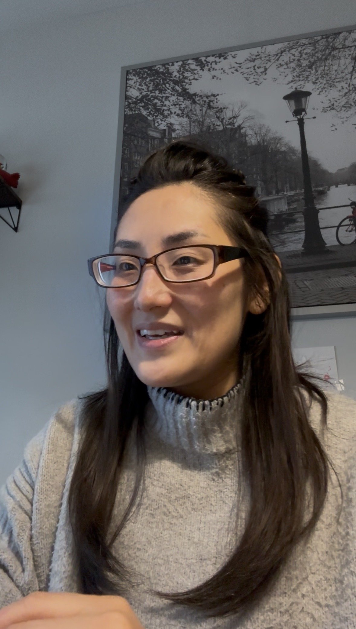 A woman with glasses smiling, wearing a beige sweater with a high neck, and with long brown hair, sitting indoors, with a black-and-white cityscape painting behind her, who is a founder of SOEL counselling.