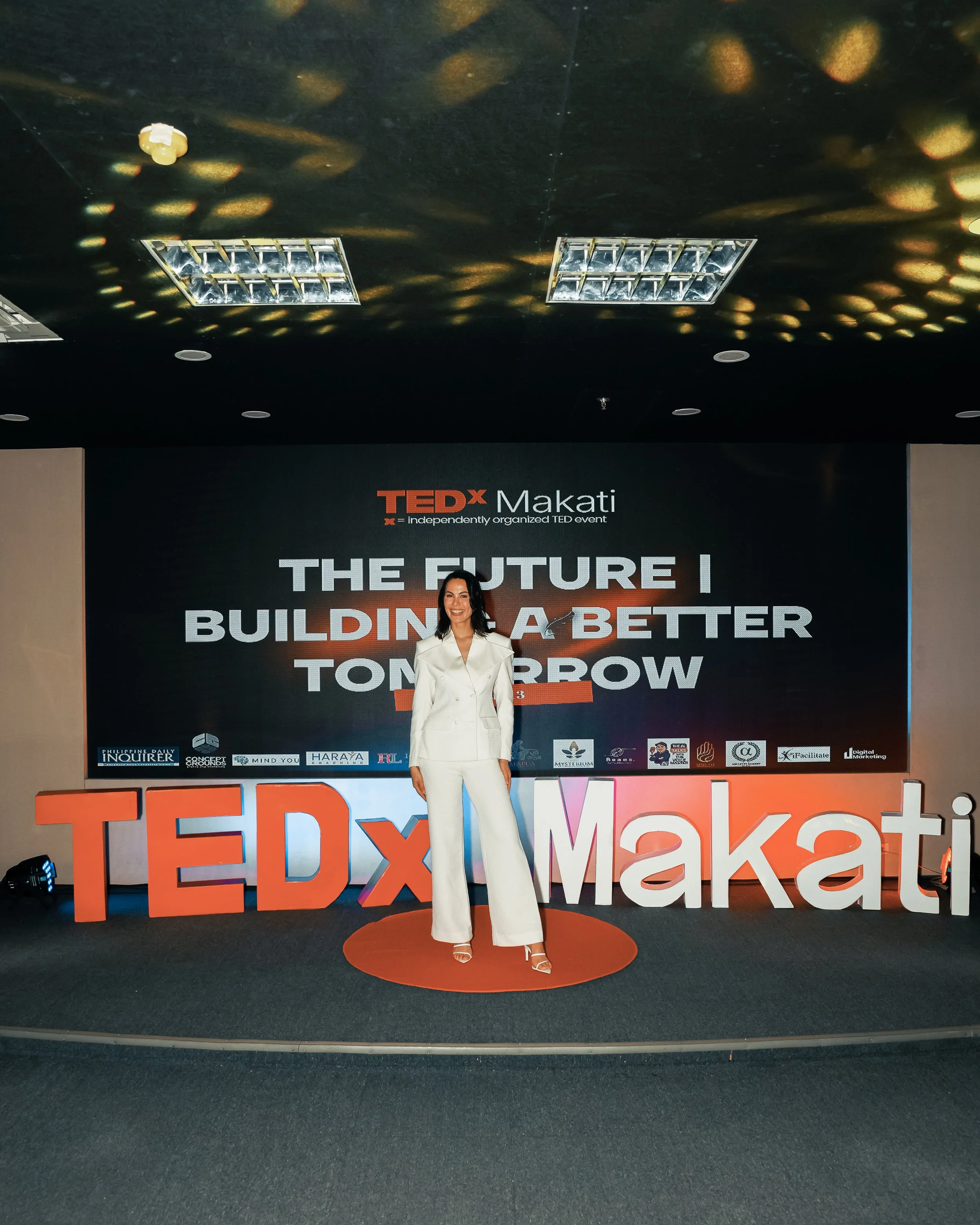 A woman in white pants and a matching white blazer stands on an orange carpeted circle in front of a large sign that reads "TEDx Makati." The background features a black screen displaying "THE FUTURE I" in large white letters, with logos of sponsors at the bottom. The ceiling has several rectangular lights and reflective surfaces.