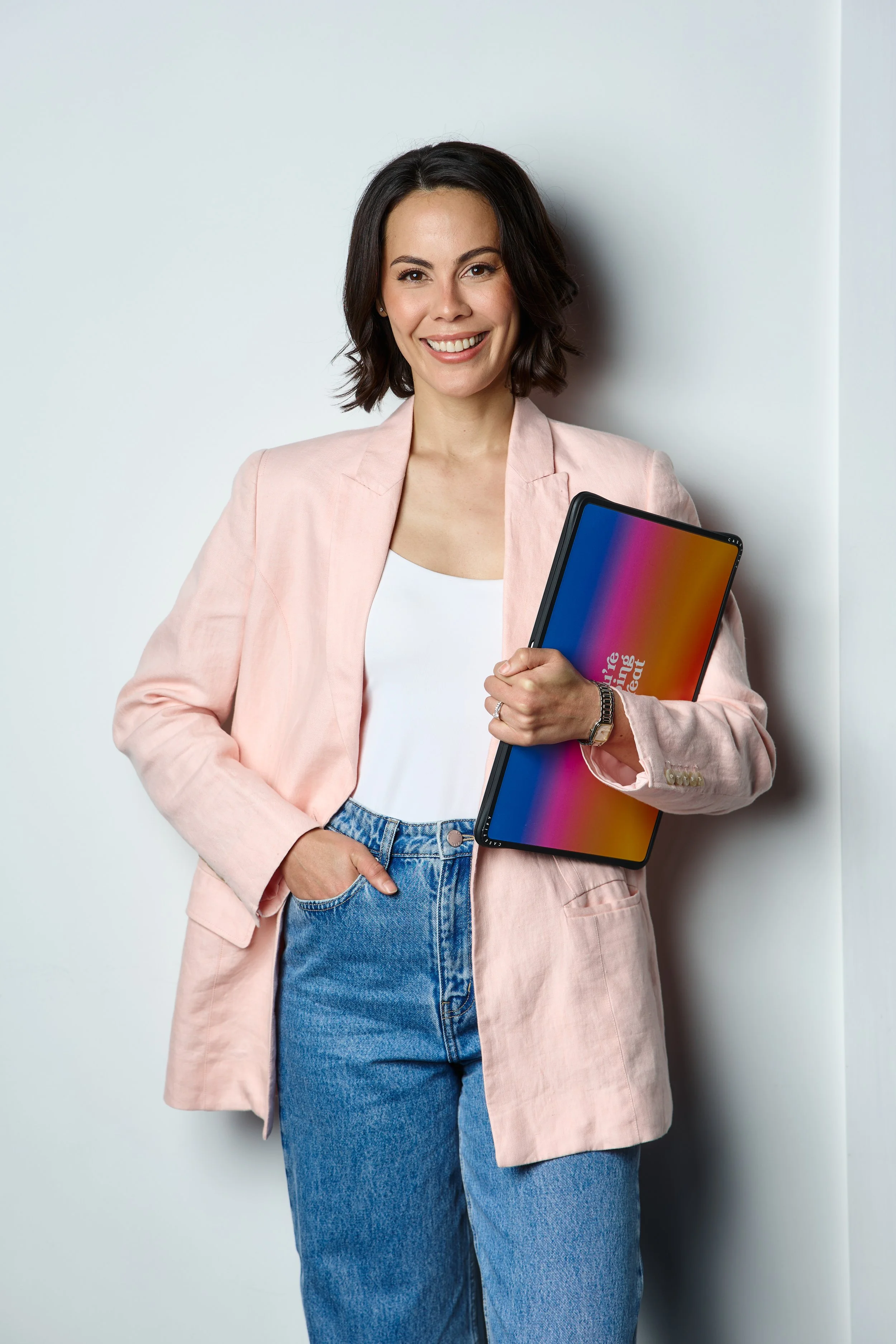 A smiling woman with shoulder-length dark hair, wearing a light pink blazer and blue jeans, holding a tablet with a colorful screen, standing against a plain white wall.