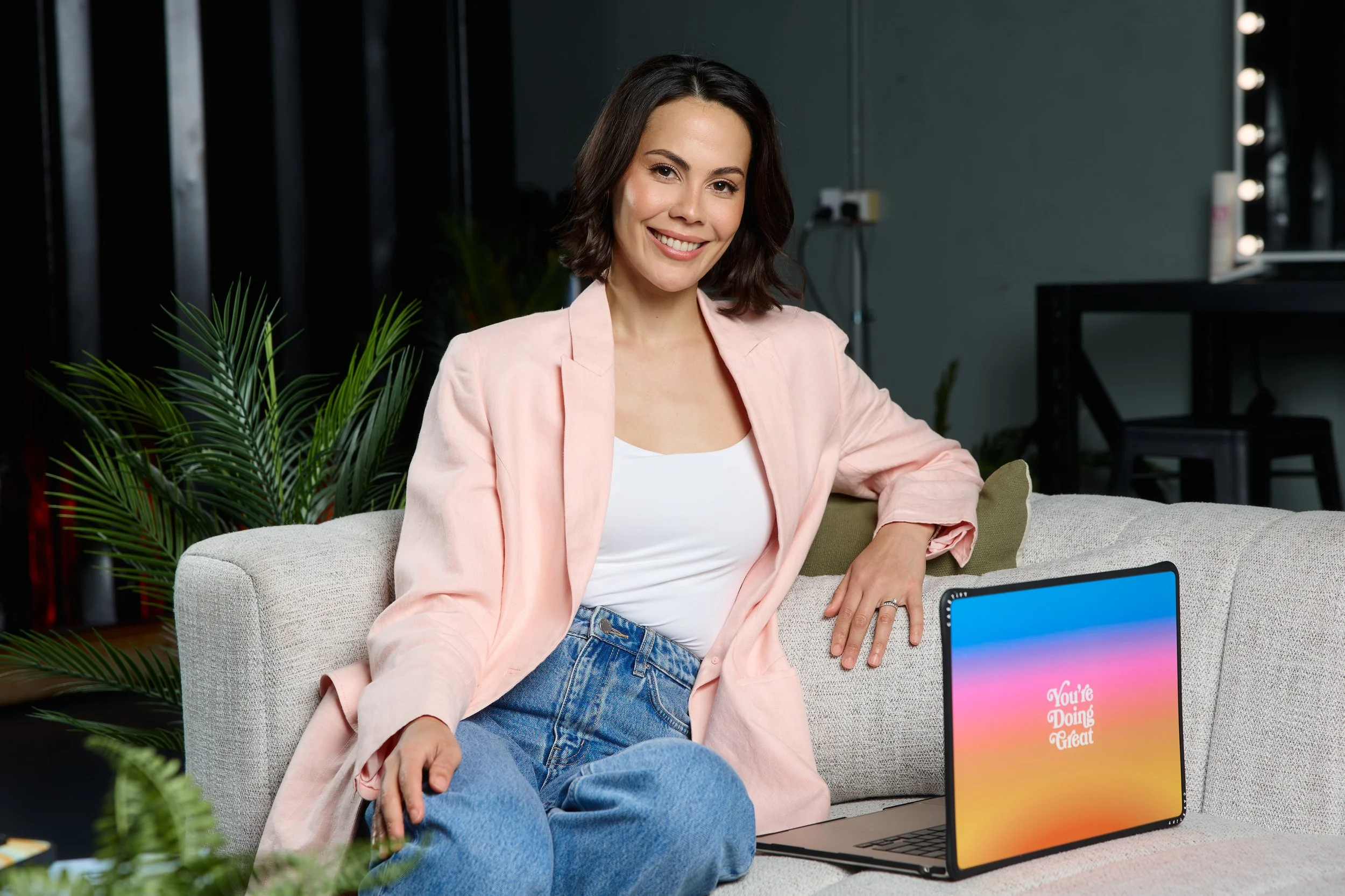 A woman with dark wavy hair wearing a light pink blazer and jeans, sitting on a beige couch in a modern office or studio setting, smiling at the camera.