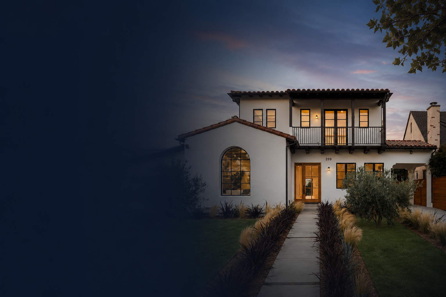 A modern two-story house at dusk with illuminated windows, a front walkway lined with dark and light plants, and a manicured lawn.