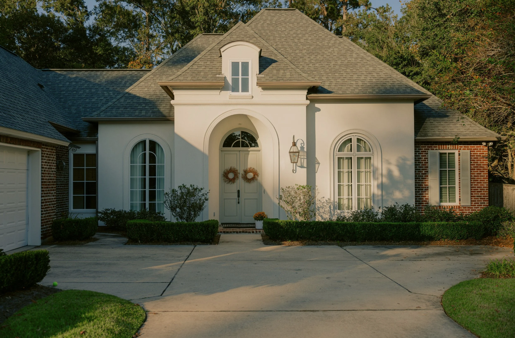 Front view of a house with a white facade, brick accents, arched windows, and a double front door decorated with wreaths. The house has a gabled roof and outdoor lighting fixtures, surrounded by a well-maintained yard with bushes and trees.