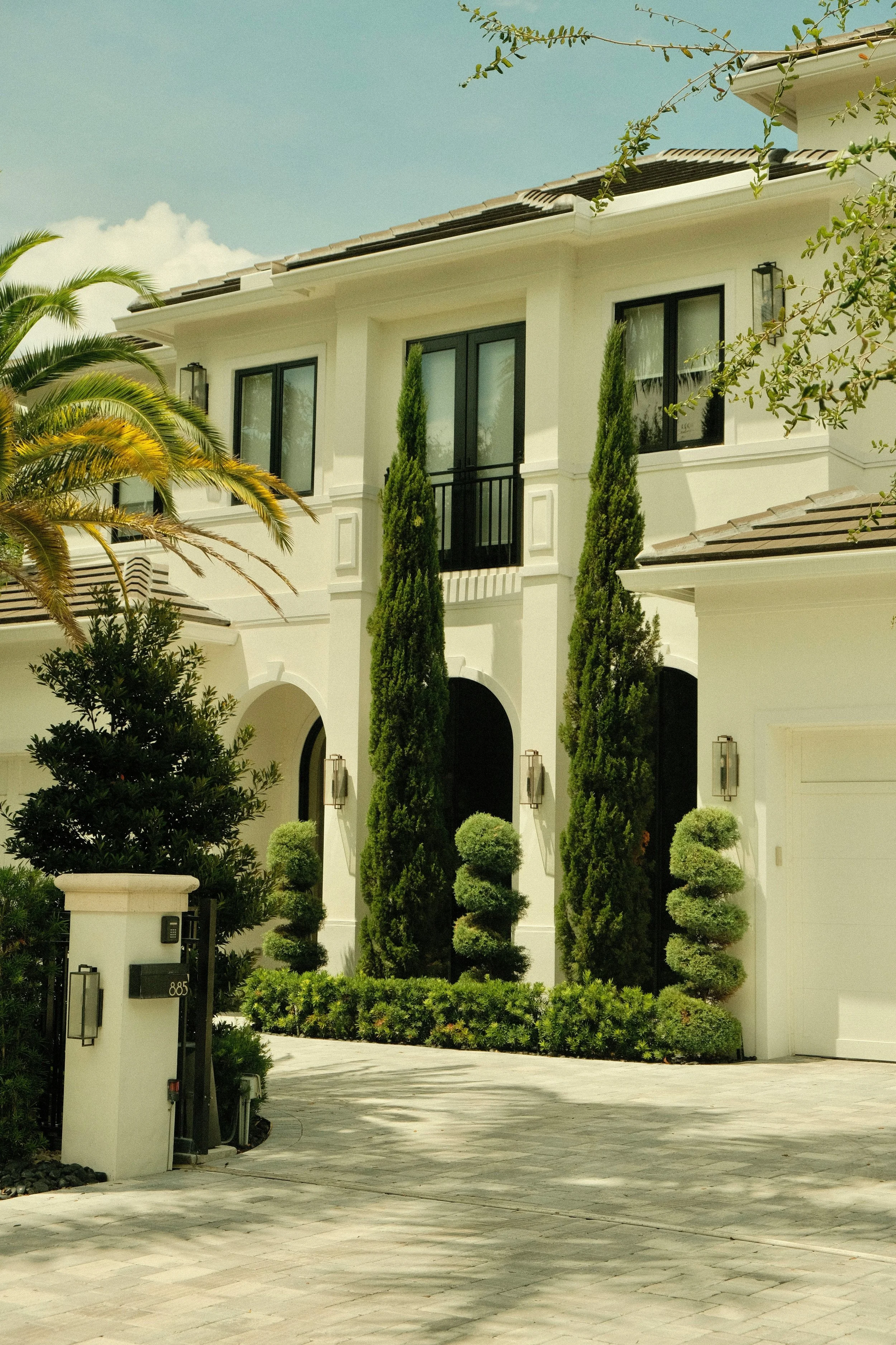 Front view of a white modern house with black window frames, tall cypress trees, and a landscaped yard with small trimmed bushes and palm trees.