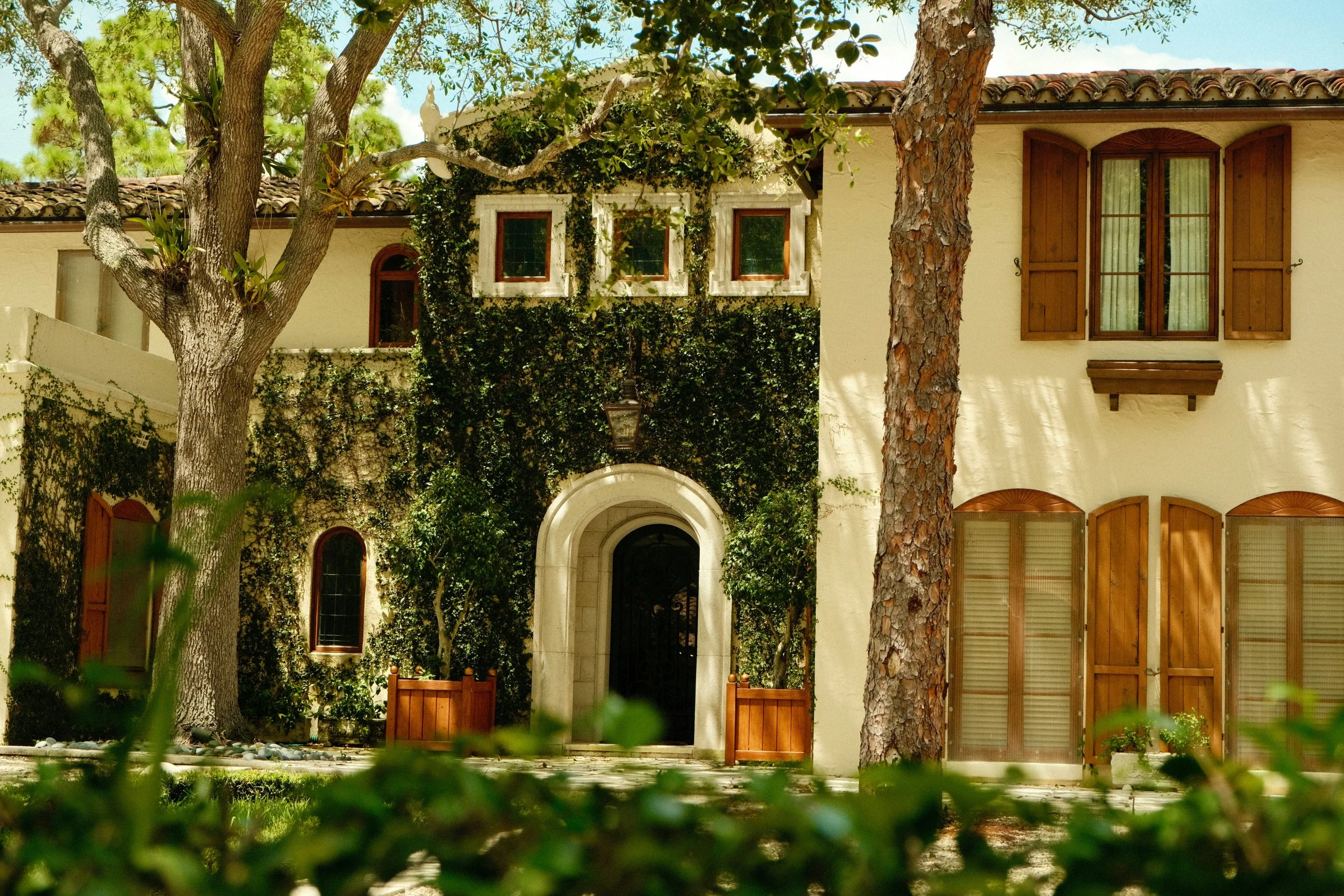 View of a house with a beige stucco exterior, brown wooden shutters, and a tiled roof, surrounded by trees and greenery.