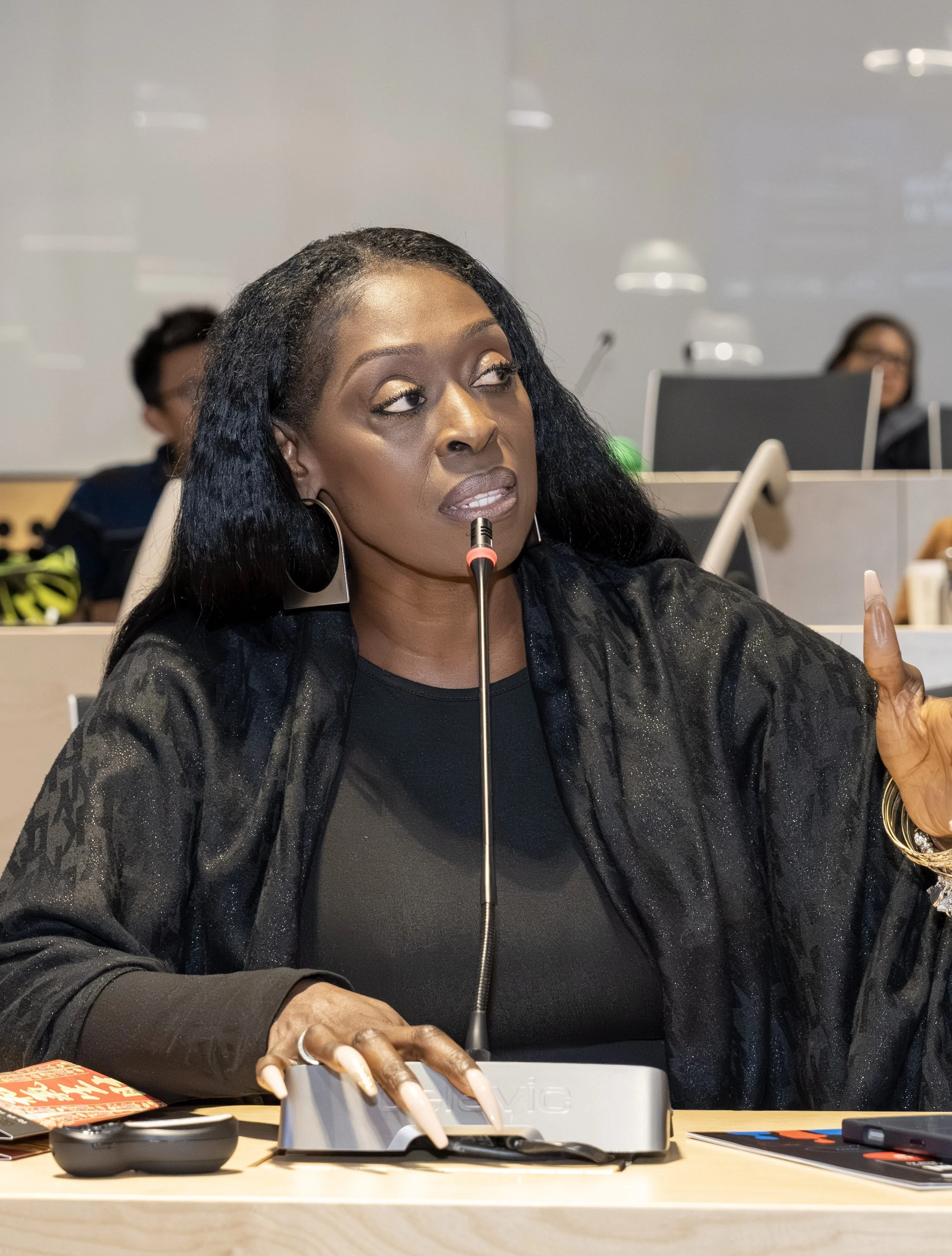 A woman with long black hair, gold eyeshadow, and earrings, sits at a desk in a meeting room, speaking into a microphone, with other people and laptops in the background.