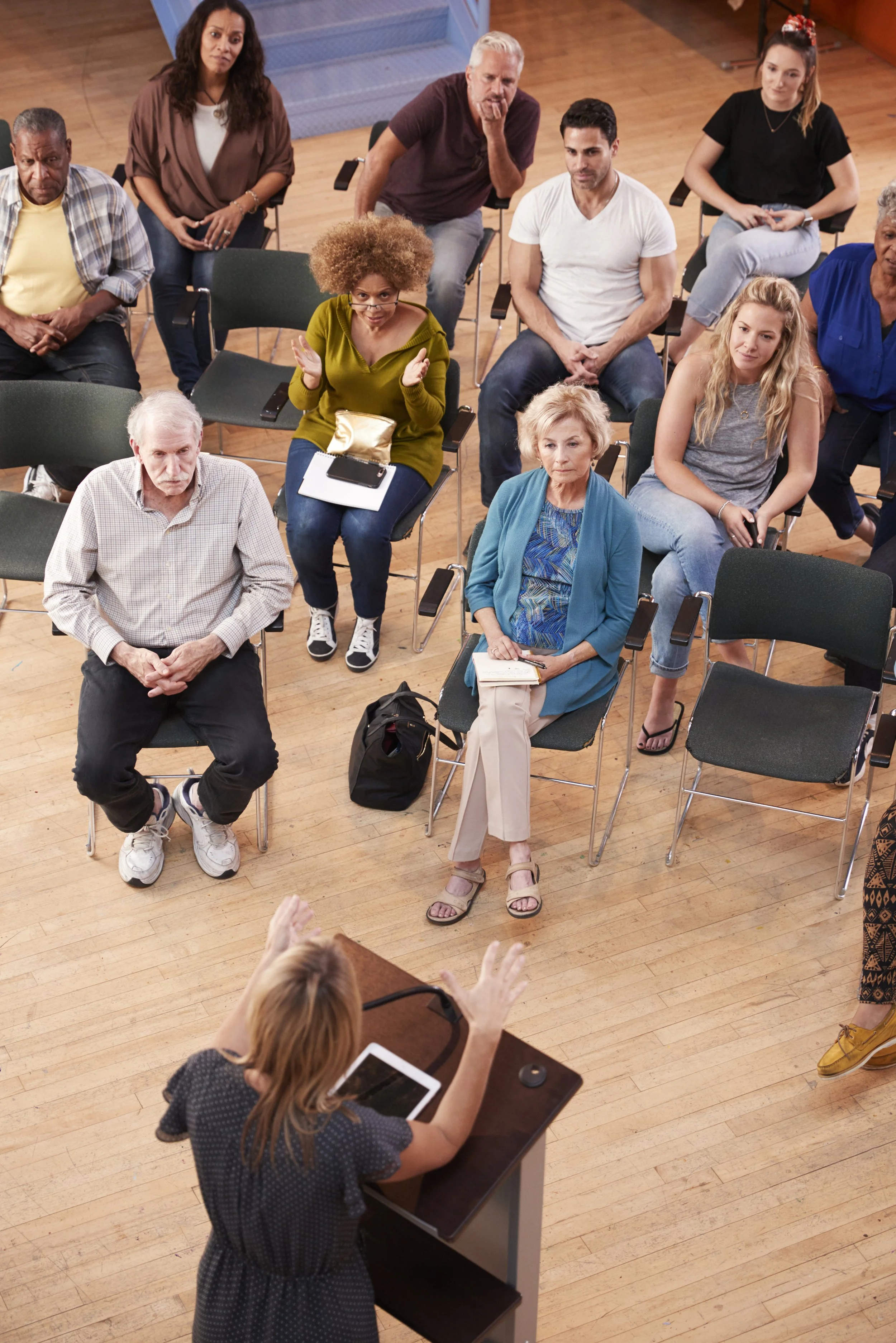 A diverse group of people seated in a classroom, some listening attentively, others appearing thoughtful, with a woman in the foreground speaking and gesturing towards the group.