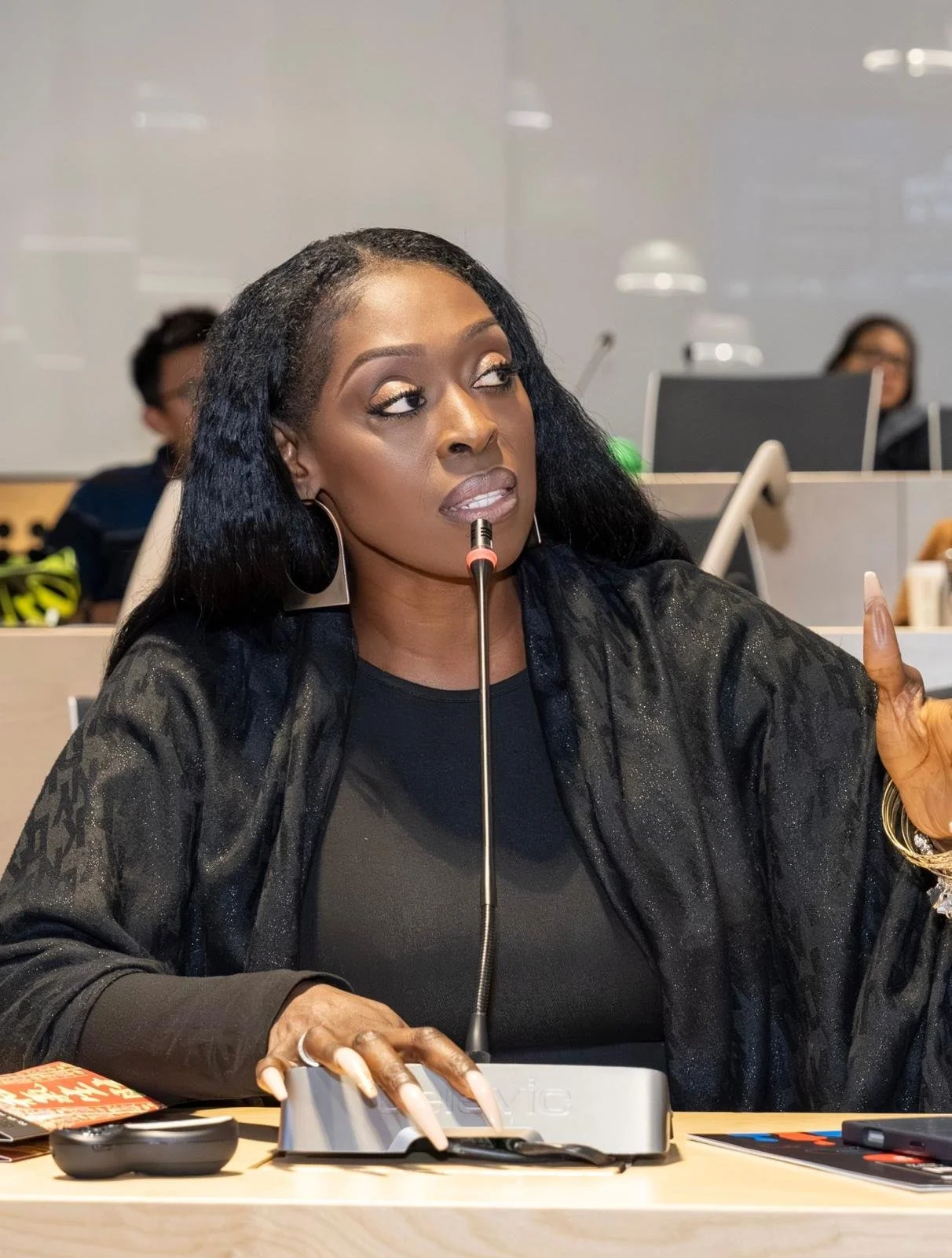 A woman with long black hair and large earrings, speaking into a microphone at a conference table with a computer and other items.