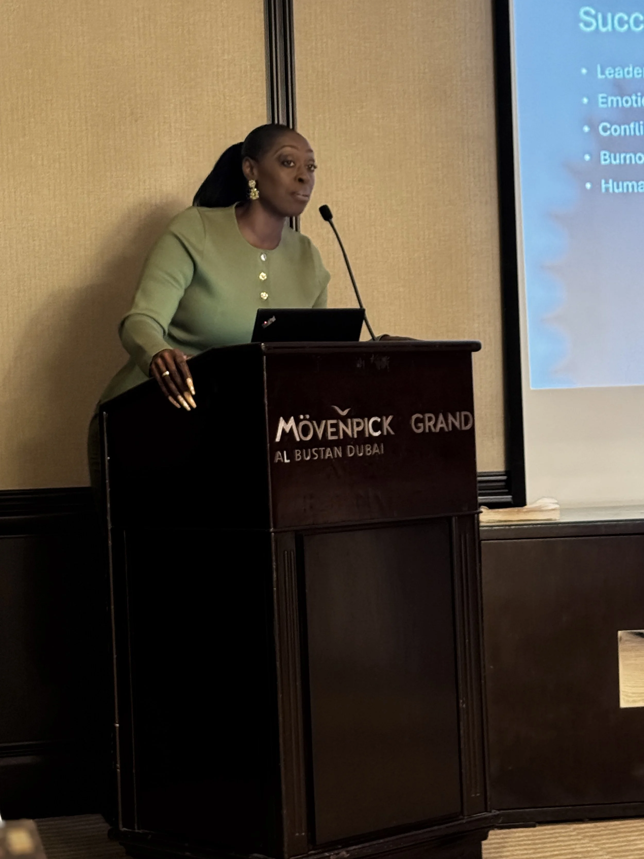 A woman standing at a podium giving a presentation, with a large screen to her right displaying a PowerPoint slide.