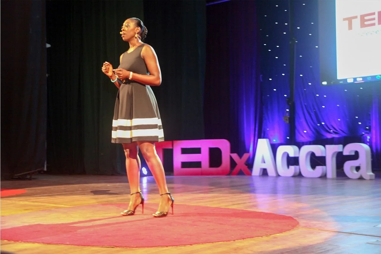 Woman in black dress giving a talk on stage at TEDxAccra event.
