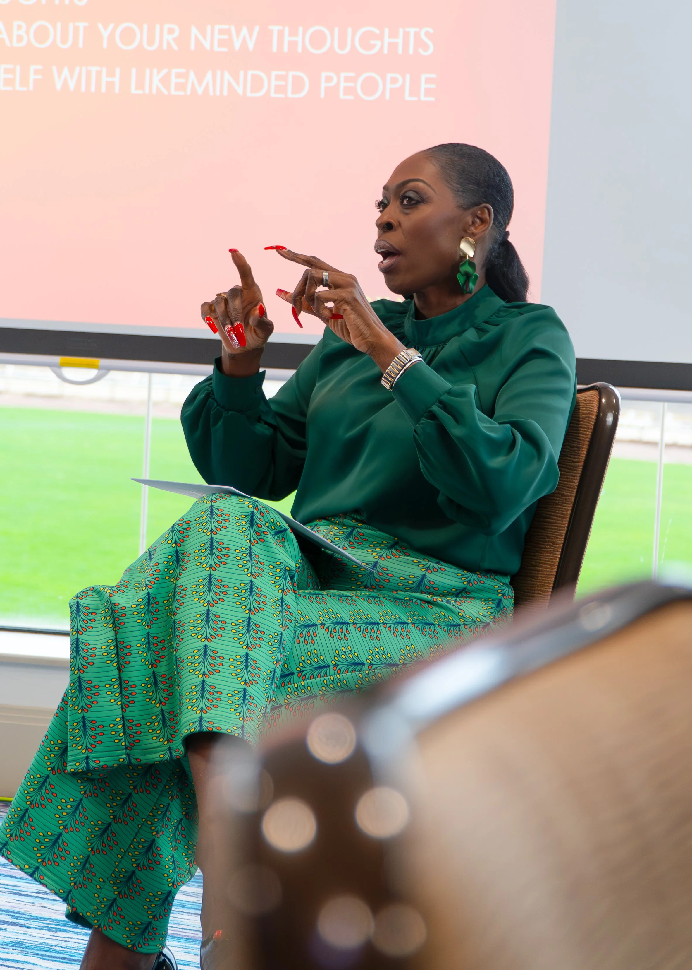 Person seated in a room with a presentation screen, wearing a green top and a patterned skirt, gesturing with hands, with a blurred object in the foreground.