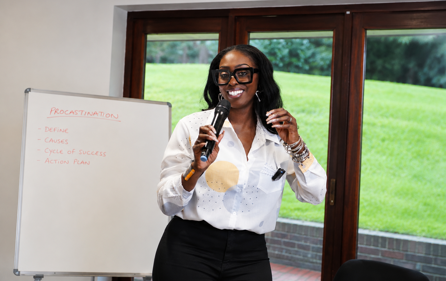 A woman with dark hair, large glasses, and jewelry, speaking into a microphone in front of a whiteboard with notes about procrastination, inside a room with large windows showing a grassy outdoor landscape.