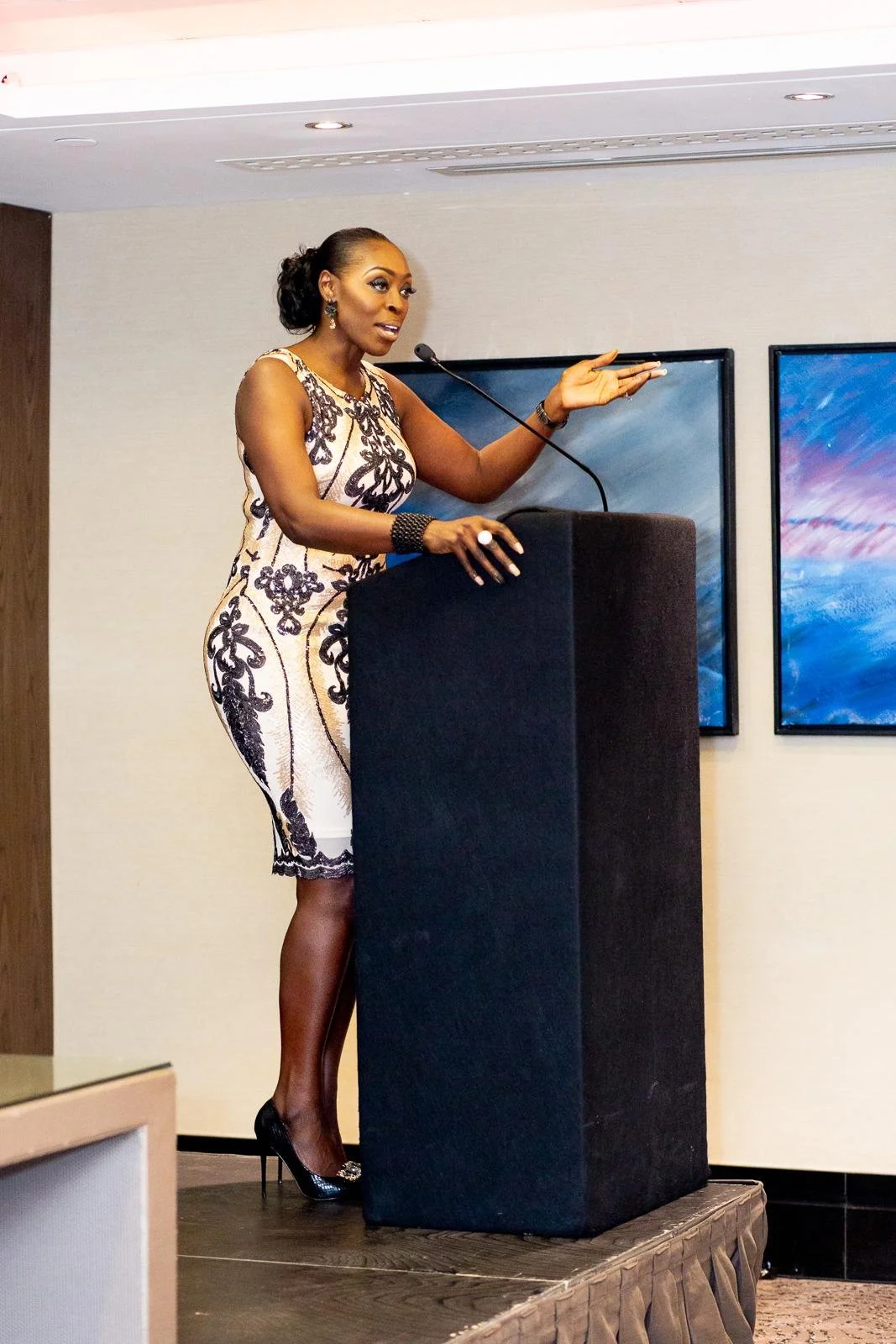 A woman in a sleeveless black and white patterned dress is speaking at a podium in a conference room, gesturing with her right hand and looking engaged.
