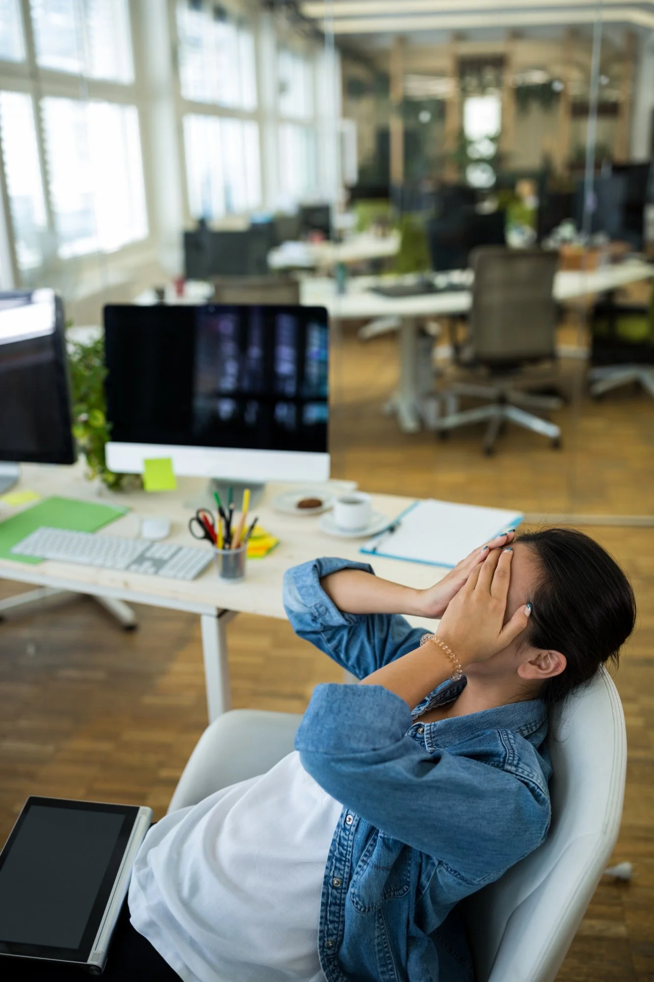 A woman sitting in an office chair, holding her head with both hands in front of a desk with a tablet and office supplies, in a modern open-plan office environment.