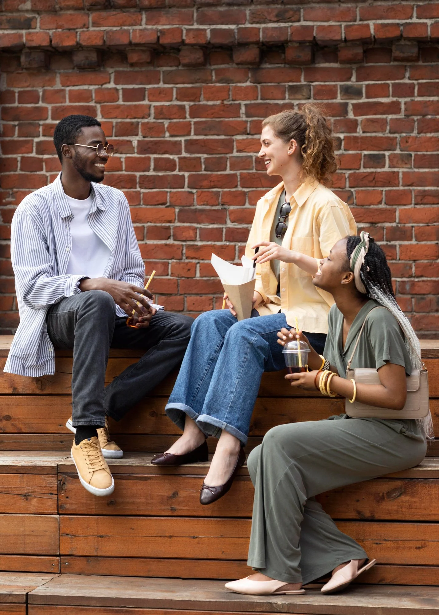 Three young women and one young man sitting on wooden steps against a red brick wall, engaging in conversation and smiling. One woman holds a drink, another has sunglasses hanging from her shirt, and the man is holding a drink and a snack.
