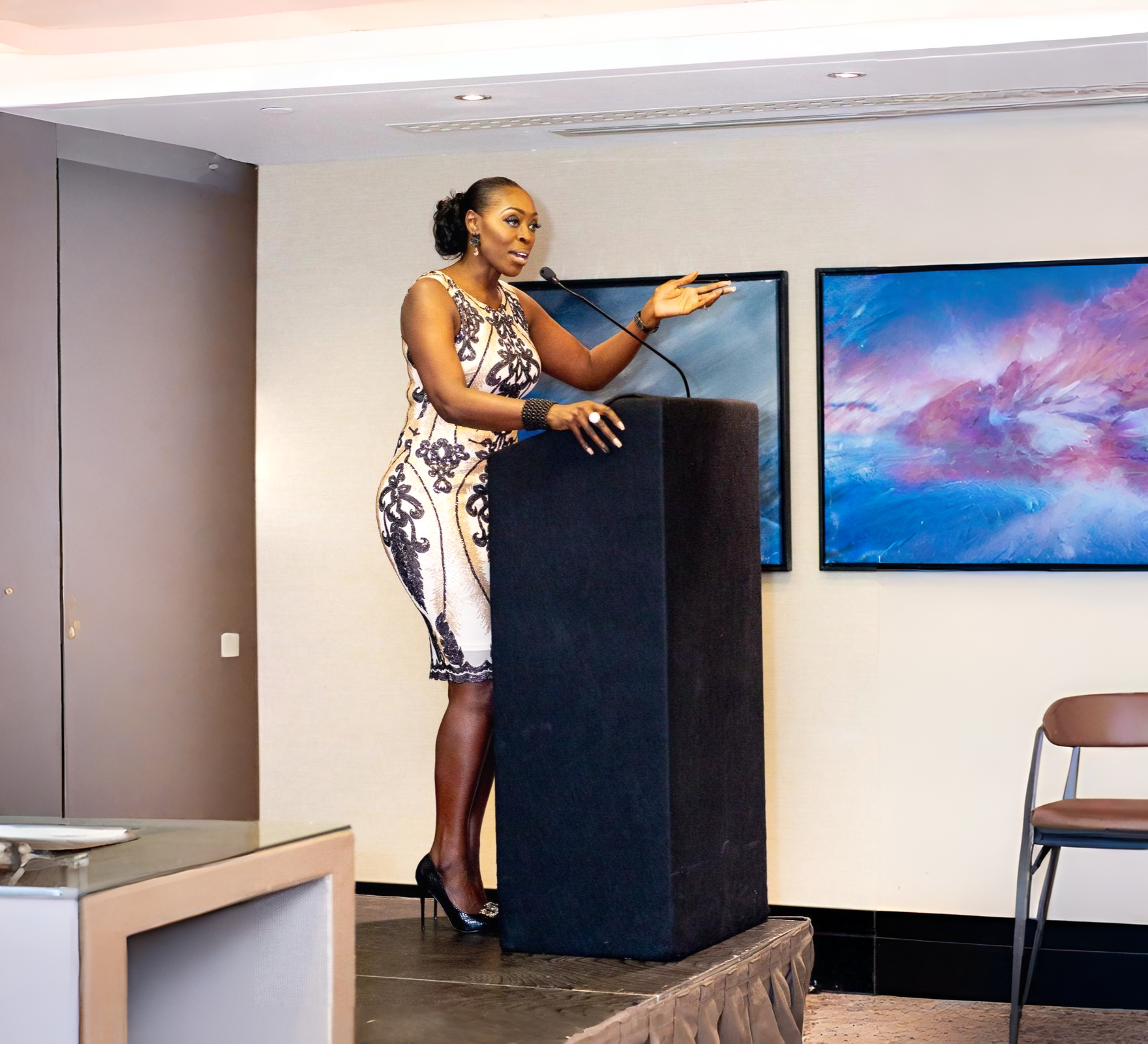 A woman in a white and black patterned dress speaking at a podium in a conference room with two abstract paintings on the wall behind her.
