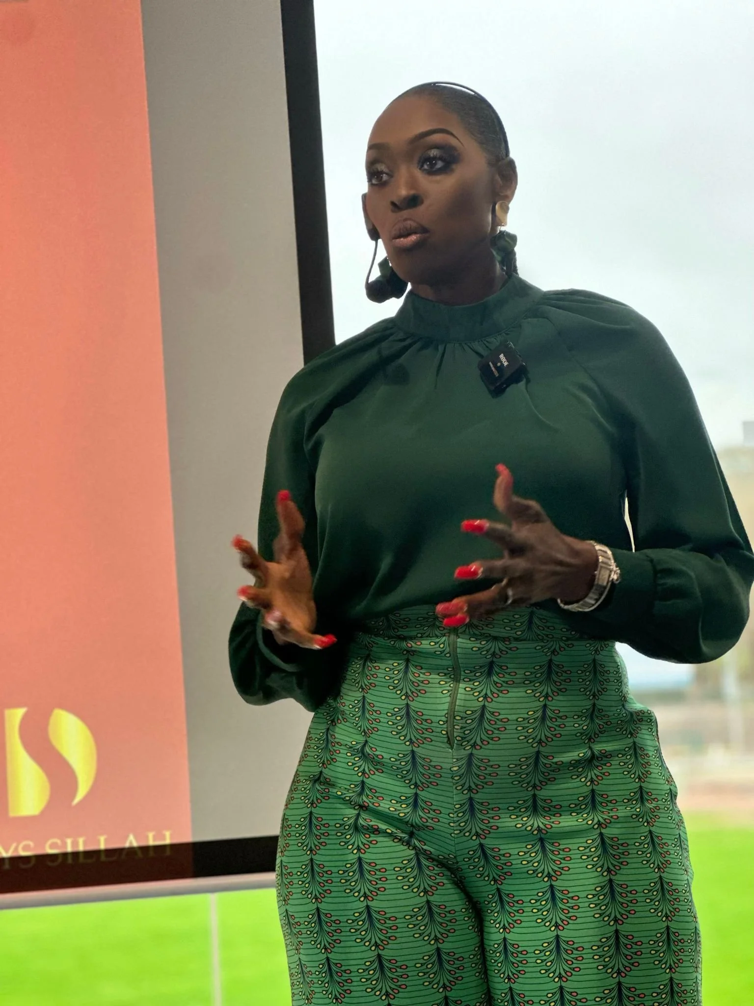A woman wearing a green patterned outfit and earrings stands in front of a presentation screen. She has dark hair pulled back and is speaking with her hands.