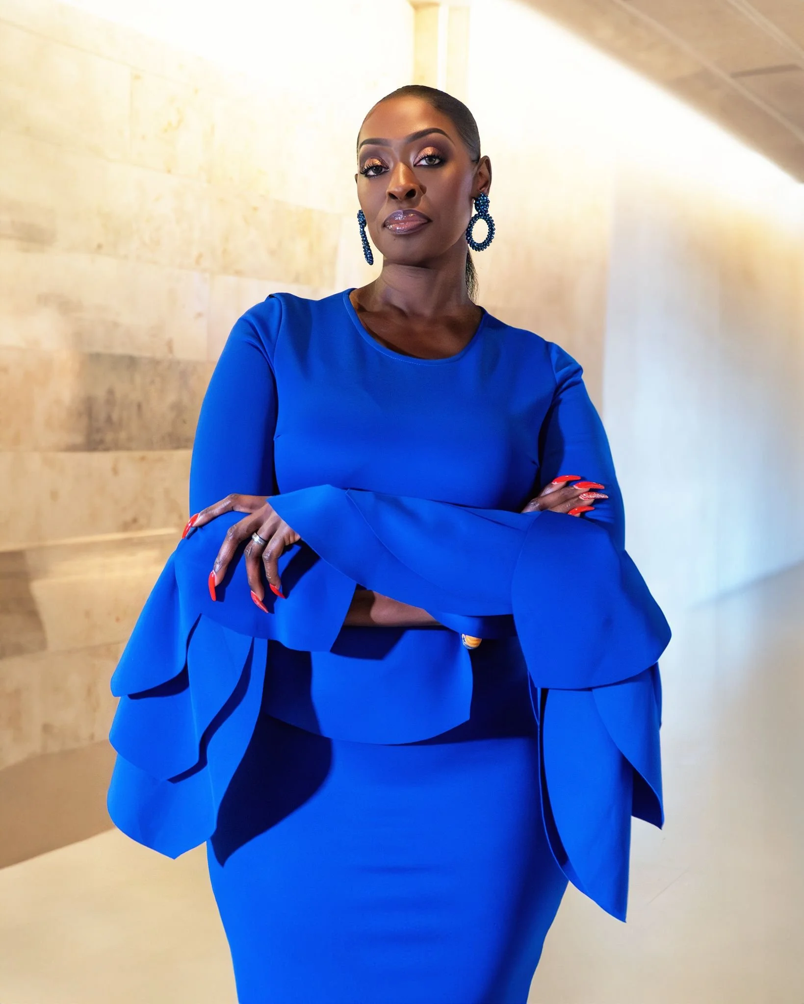 A woman dressed in a bright blue outfit with matching blue beaded earrings, standing in a well-lit indoor space with beige walls, looking confidently at the camera with crossed arms.