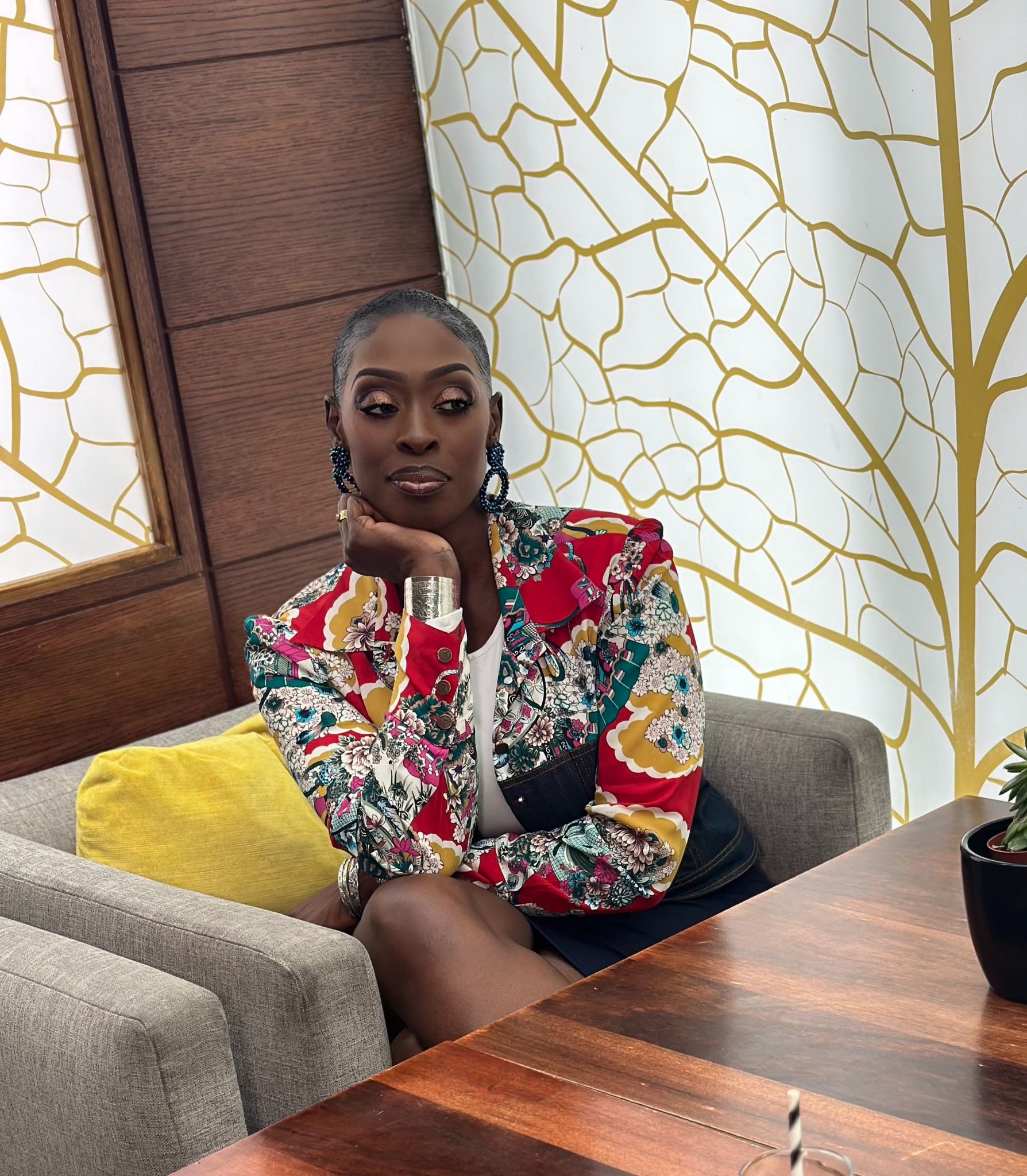 A woman with short gray hair, wearing a colorful patterned jacket with floral and geometric designs, sitting on a gray couch with a yellow pillow, in a room with a wooden wall and a gold and white decorative wall pattern.