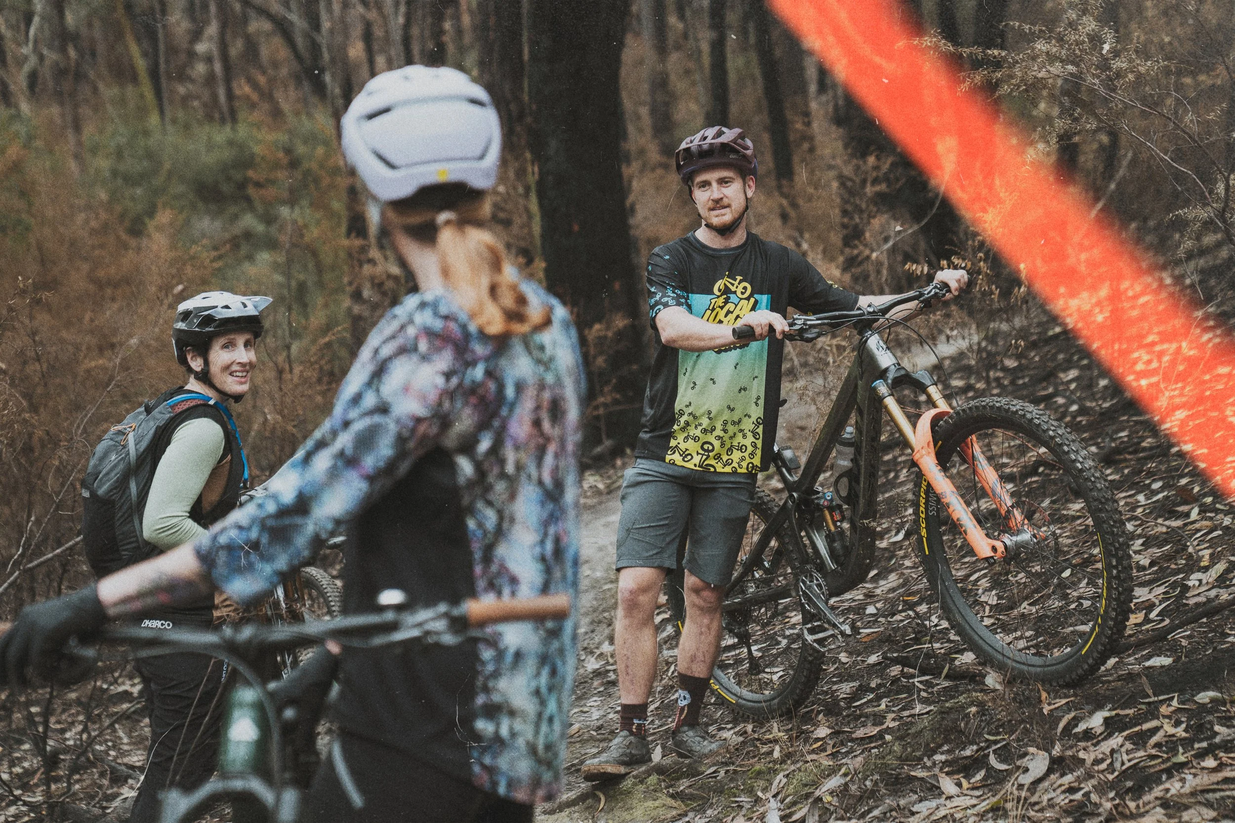 Two women and a man with mountain bikes in a wooded forest, wearing helmets and casual clothes, engaged in conversation, with autumn leaves on the ground.