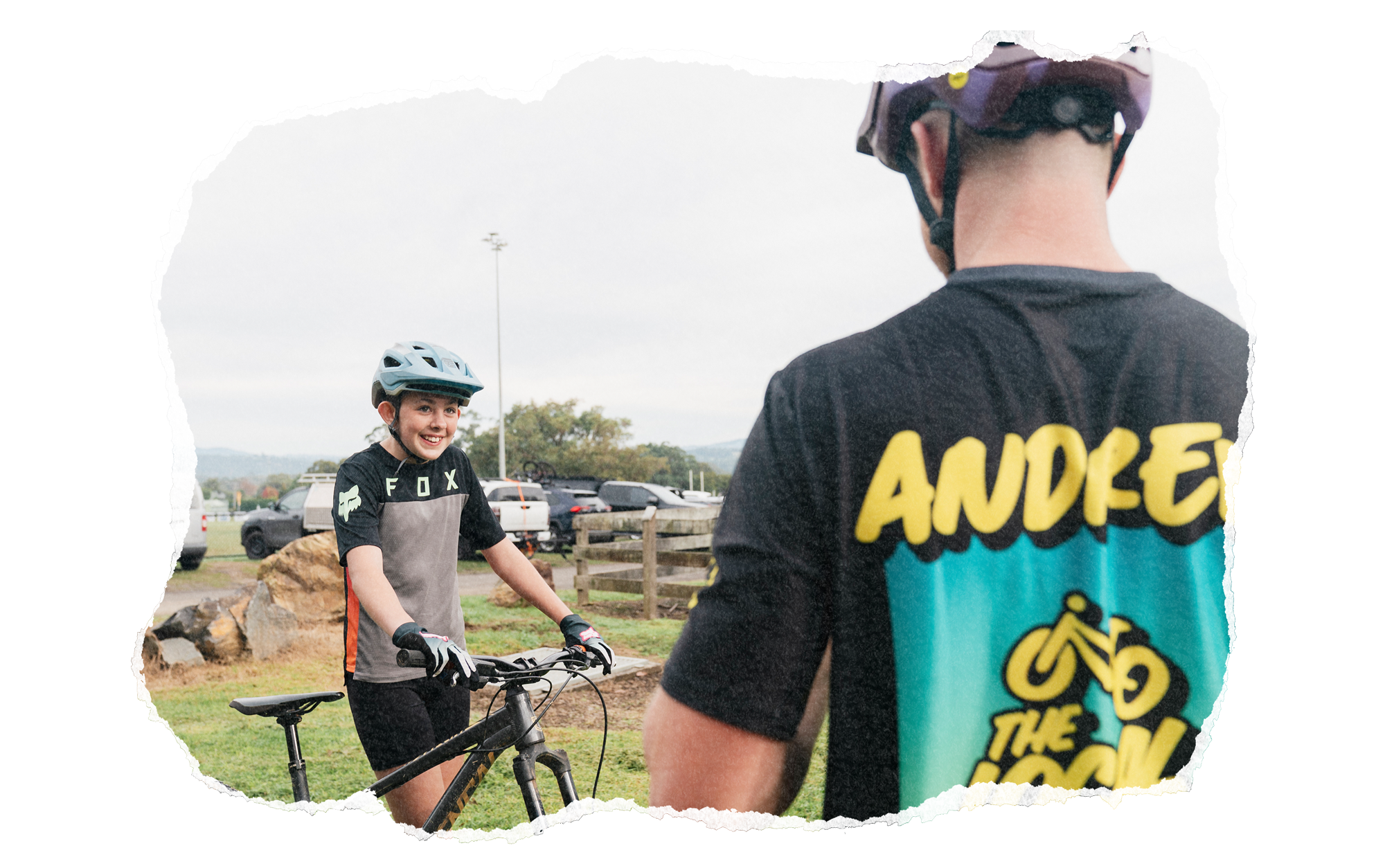 A young girl smiling while standing with her mountain bike, wearing a helmet, in an outdoor park area with rocks, grass, trees, parked cars, and cloudy sky. Another person in the foreground has their back to the camera, wearing a colorful cycling shirt with yellow text.