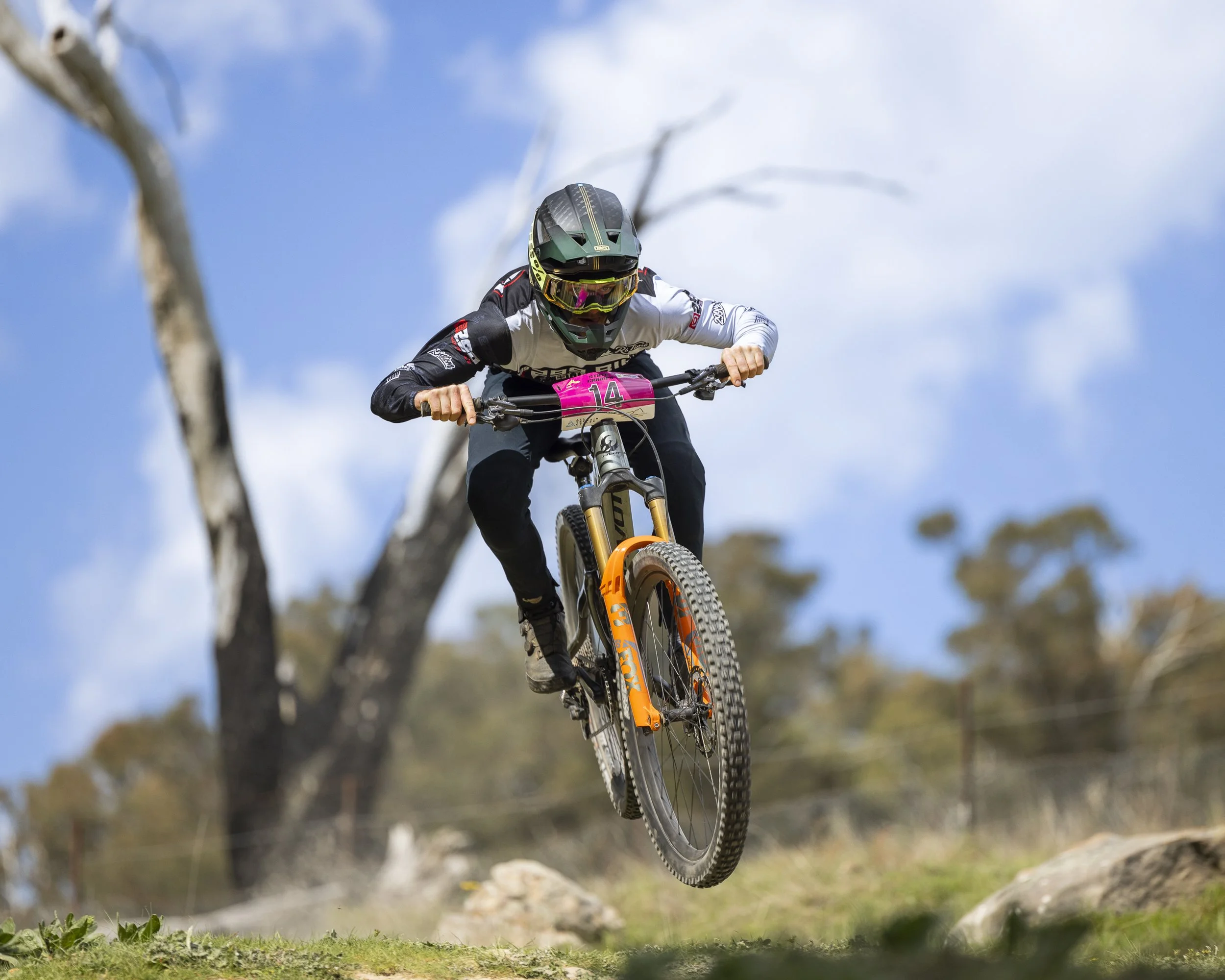A mountain biker wearing a helmet and goggles rides down a trail with trees and cloudy sky in the background.