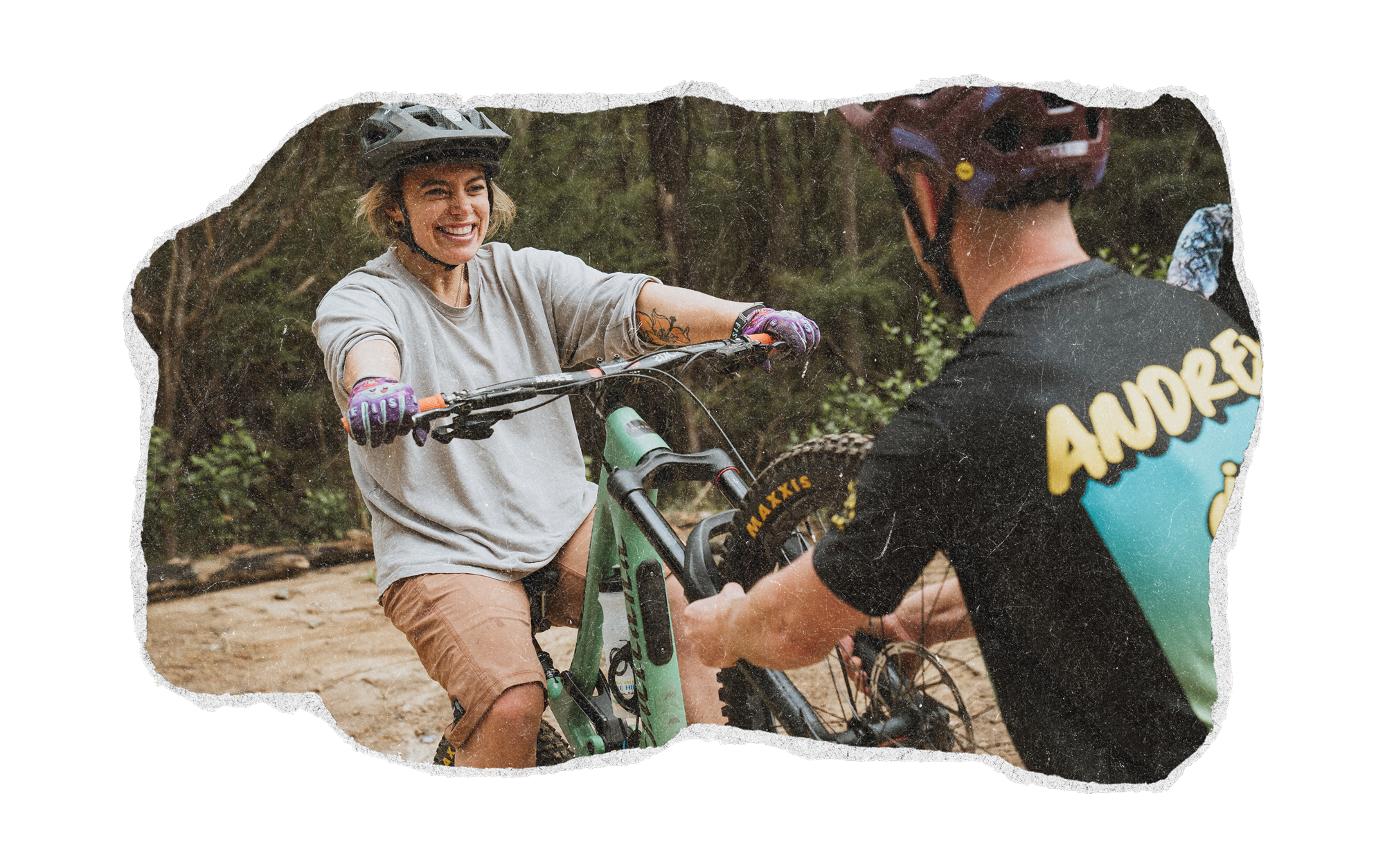 A woman smiling as she rides a mountain bike with a man helping her on a dirt trail in a wooded area, both wearing helmets.