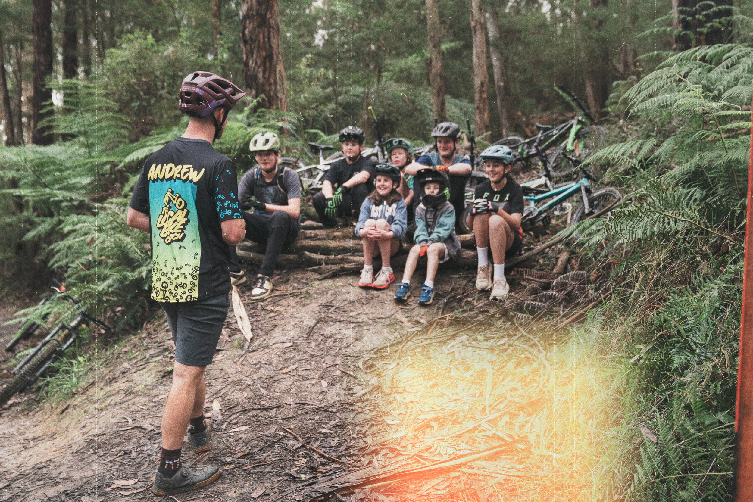 A group of children and an adult in biking helmets sitting on a log in a forest, with bicycles in the background, during a mountain biking trip.