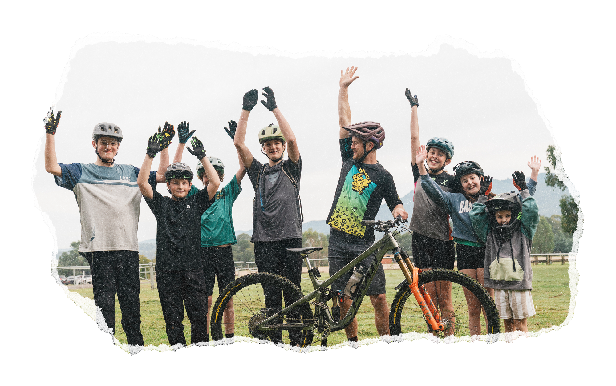 Group of kids and teenagers wearing mountain biking helmets and gloves, standing outdoors with a mountain bike, smiling and raising their hands.