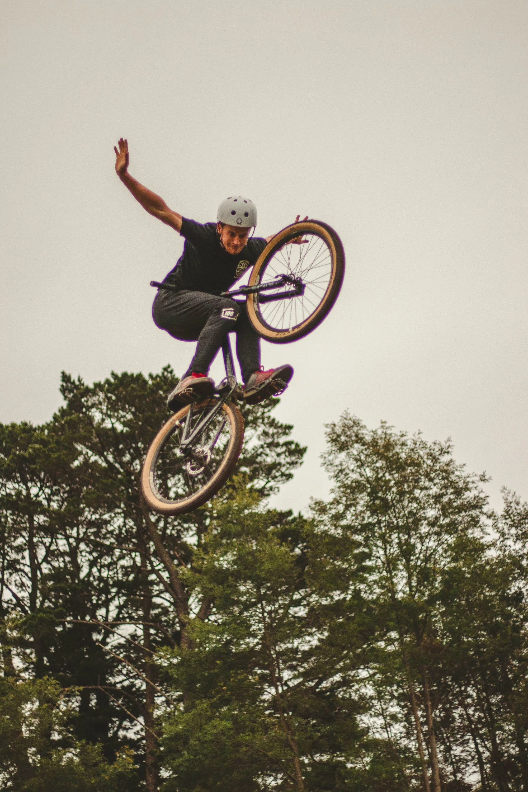 Person wearing a helmet performing a trick on a mountain bike in mid-air against a backdrop of green trees and a cloudy sky.