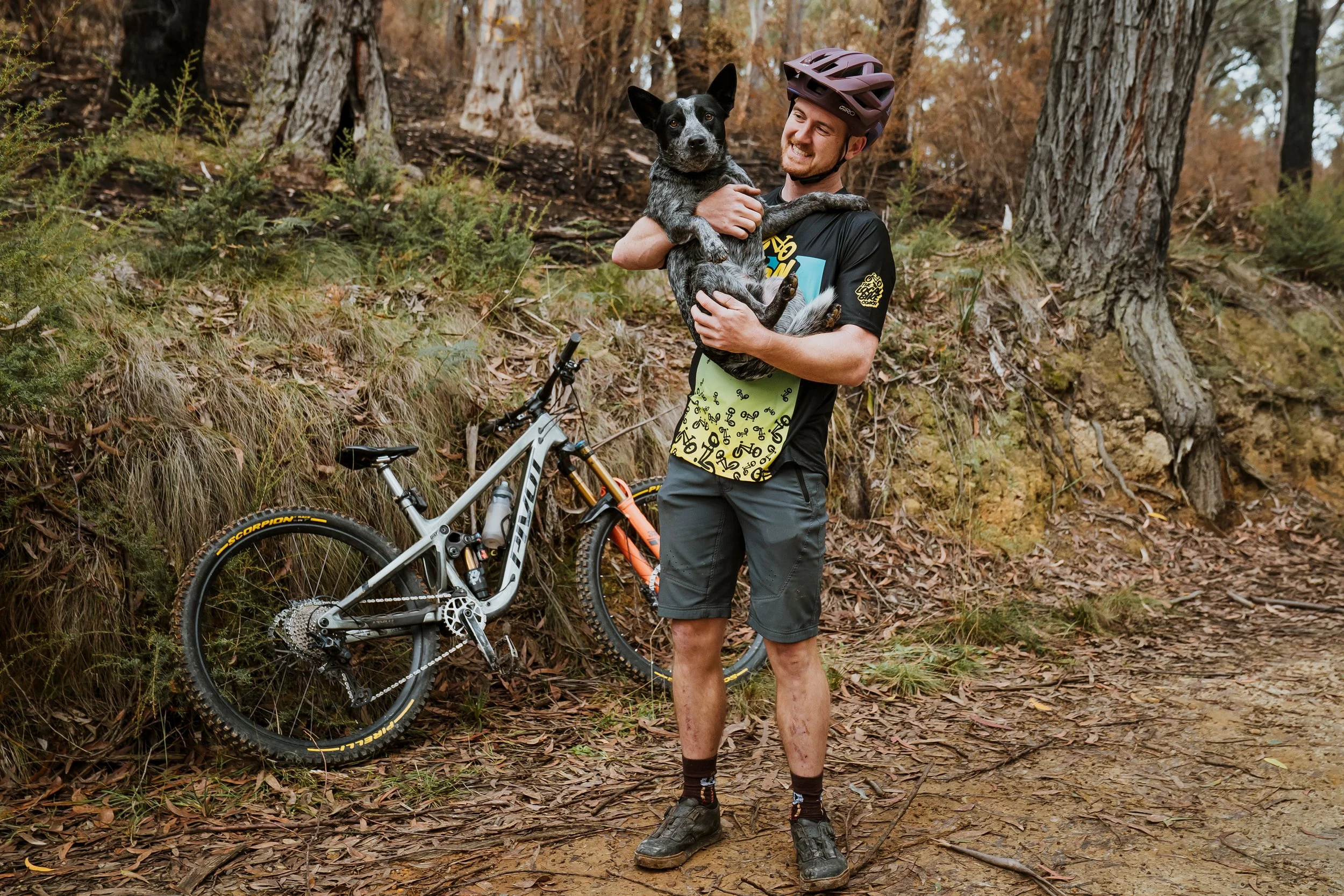 A man in cycling gear and a helmet standing on a dirt trail in a forest, holding a black and white dog, with a mountain bike leaning against a nearby tree.