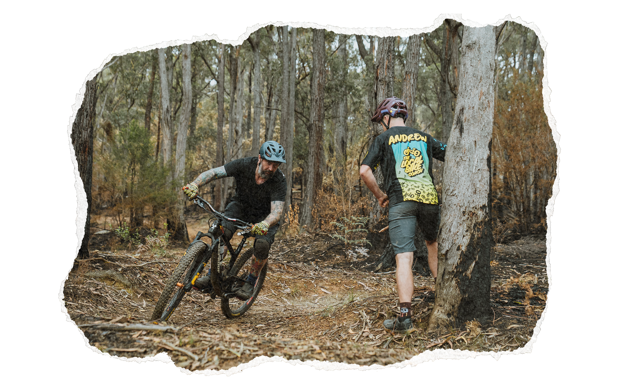 Two men in helmets and mountain biking gear are on a trail in a dense forest. One man is riding a mountain bike, and the other is standing beside a tree, watching him.