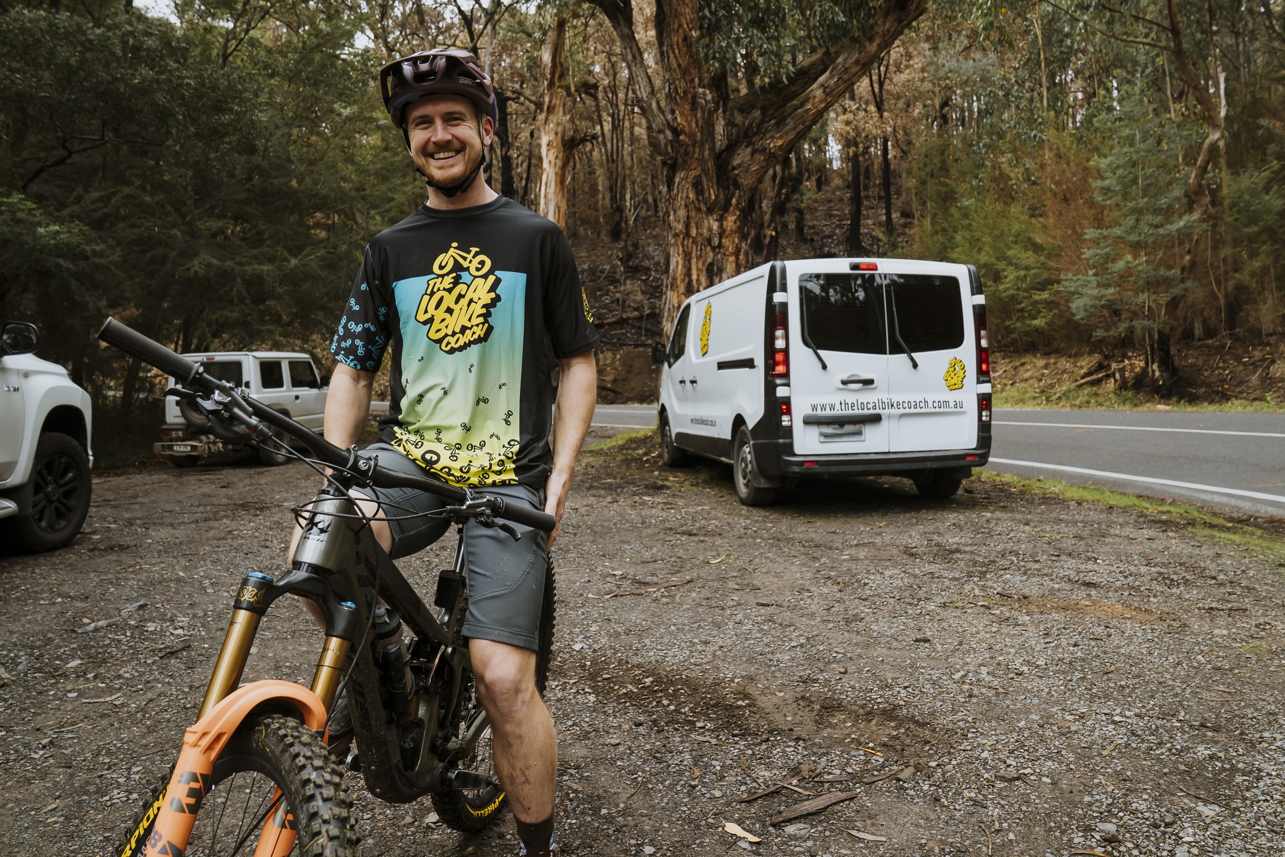 Smiling man in biking gear standing with bike in wooded area, with work vans parked on the roadside.