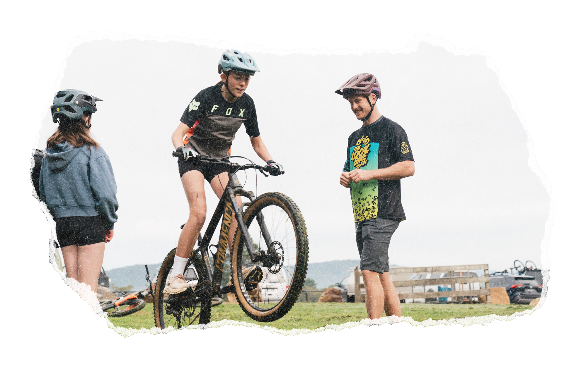 A person in a black shirt and helmet is riding a mountain bike while two others, a girl and a man, watch. The scene is outdoors on a cloudy day.